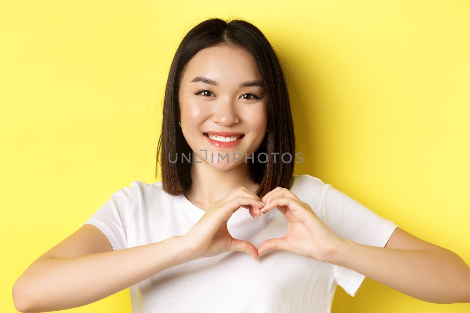 Valentines day and women concept. Close up of pretty asian girl in white t-shirt, smiling and showing heart, I love you gesture, standing over yellow background.