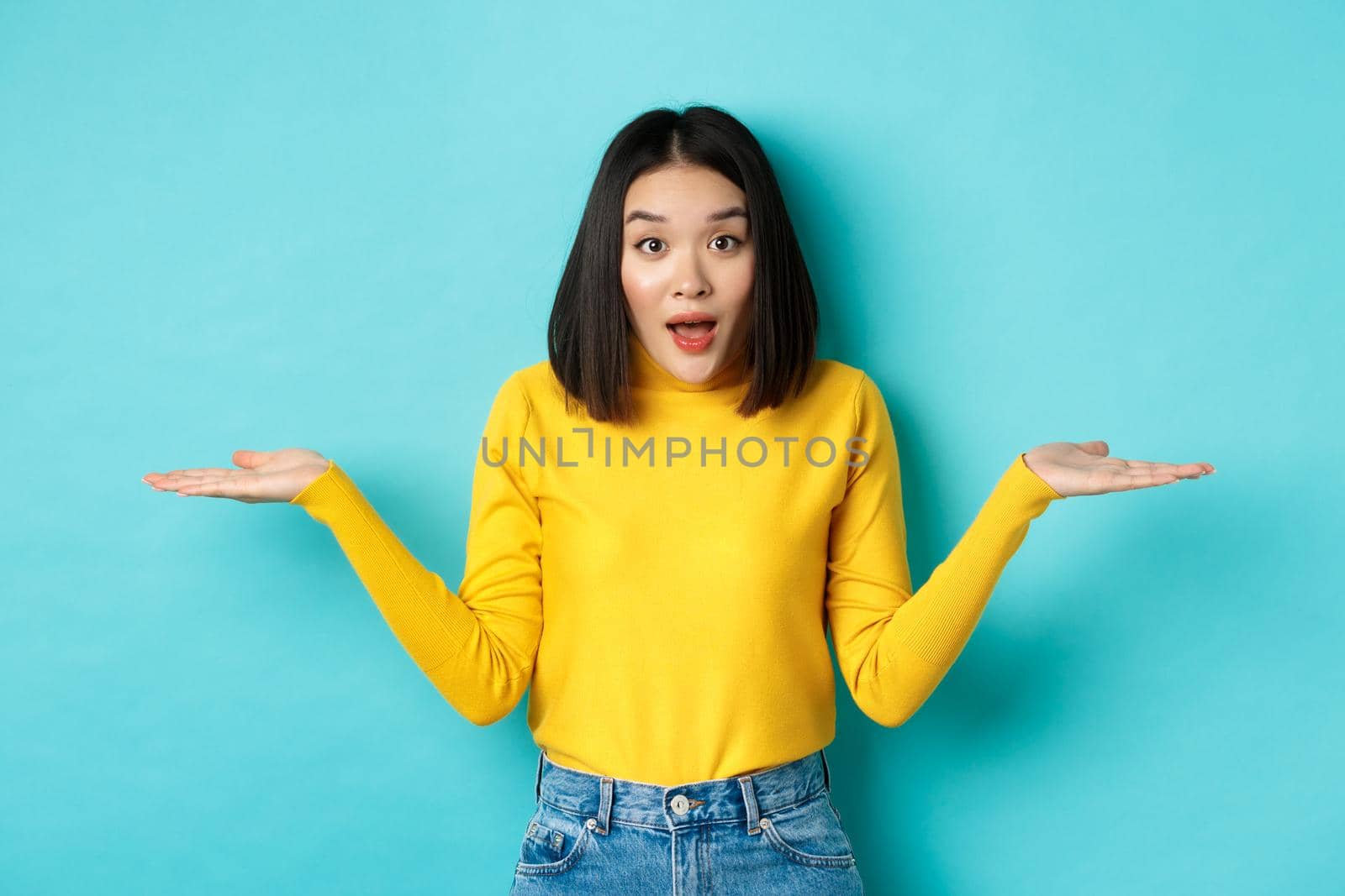Image of indecisive asian woman shrugging shoulders, spread hands sideways and looking clueless at camera, standing confused against blue background.