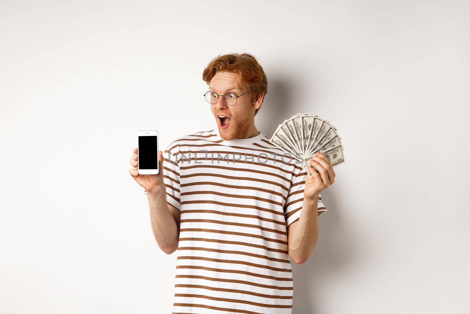 Amazed redhead man showing smartphone app on blank screen and money, winning prize cash online, standing over white background.