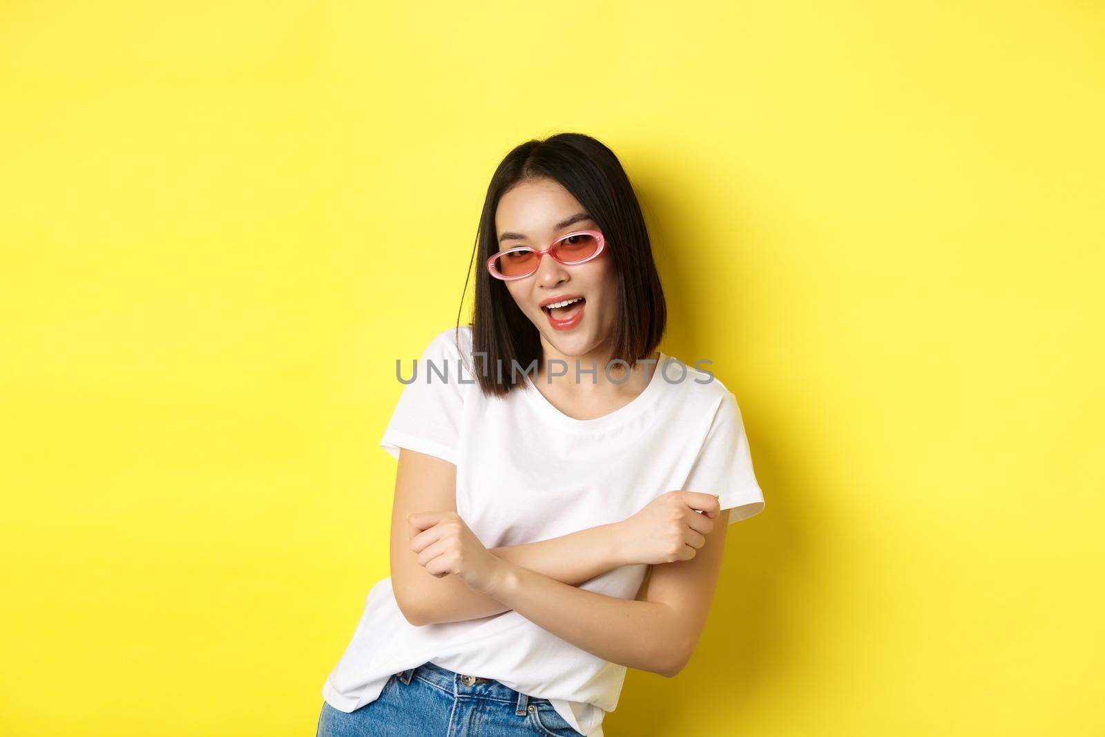 Fashion and lifestyle concept. Sassy and confident asian woman in trendy sunglasses looking self-assured at camera, standing over yellow background.