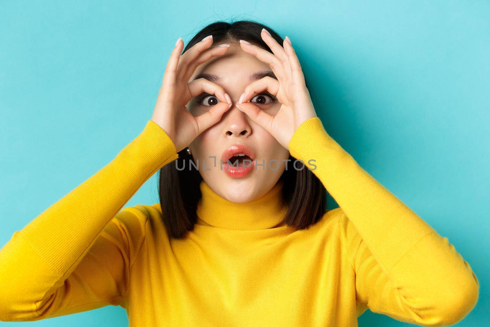Close up of funny asian woman looking through hand binoculars with surprised face, see something amazing, standing over blue background.