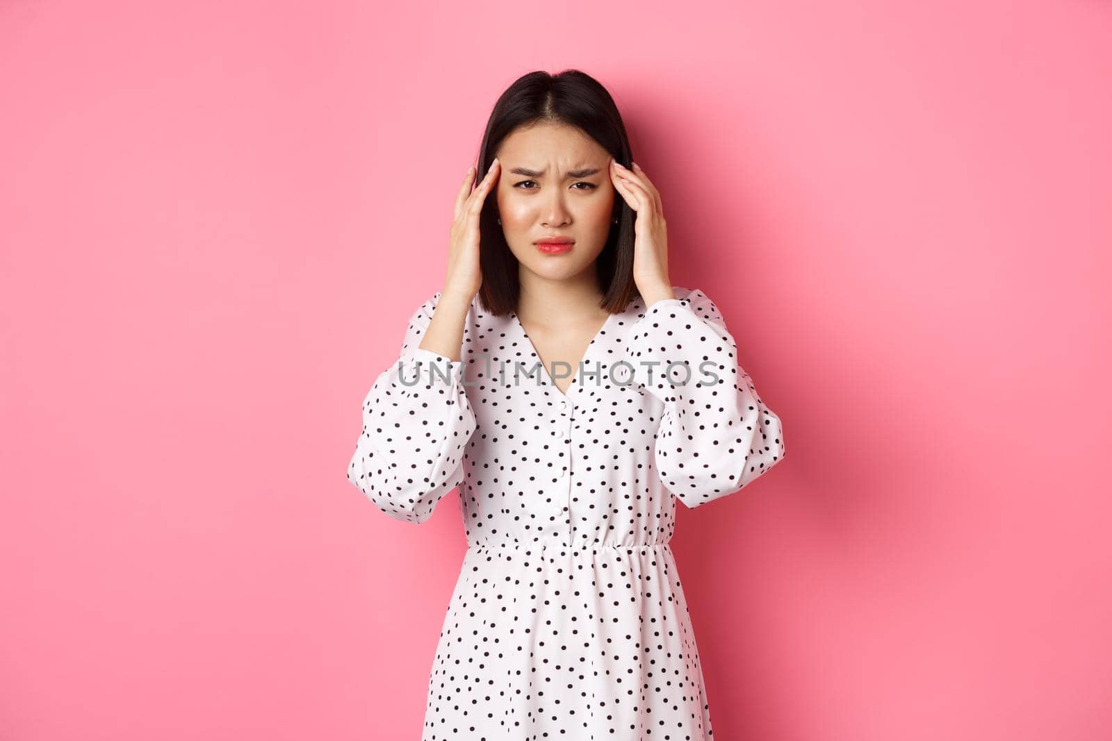 Image of upset asian woman having headache, feeling unwell or dizzy, close eyes and massaging head, suffering migraine, standing over pink background by Benzoix