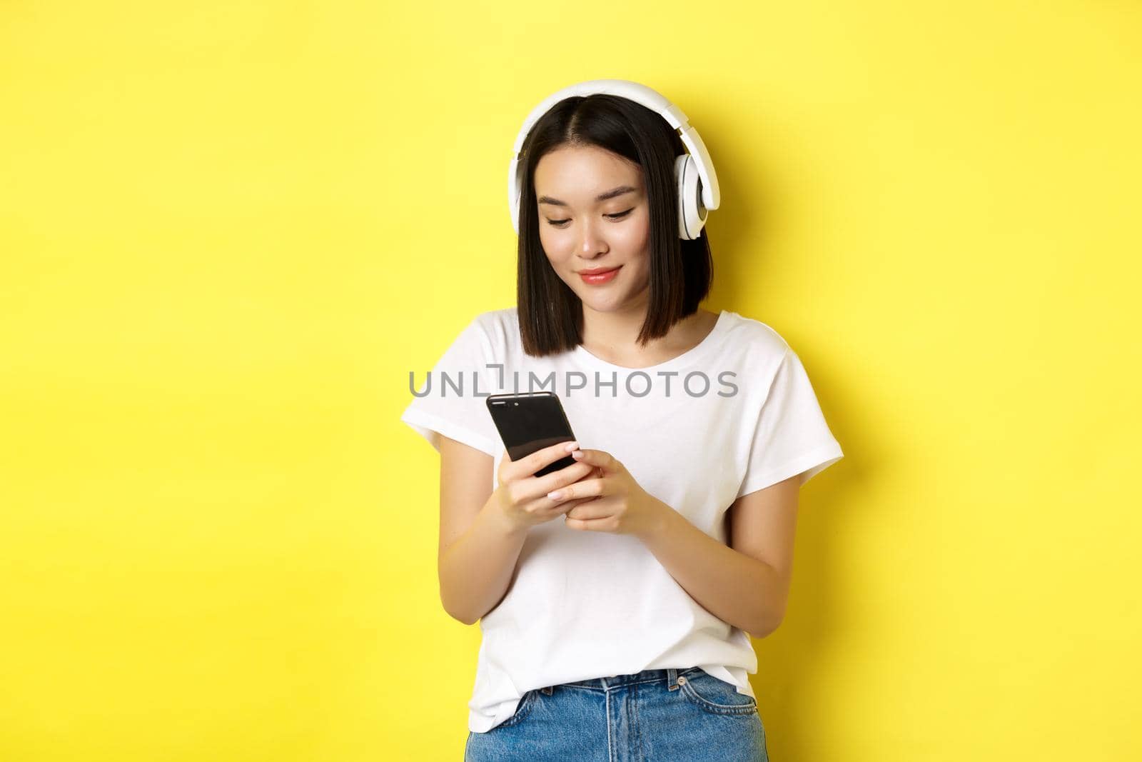 Modern asian girl listening music in wireless headphones, reading smartphone screen and smiling, standing in white t-shirt over yellow background.