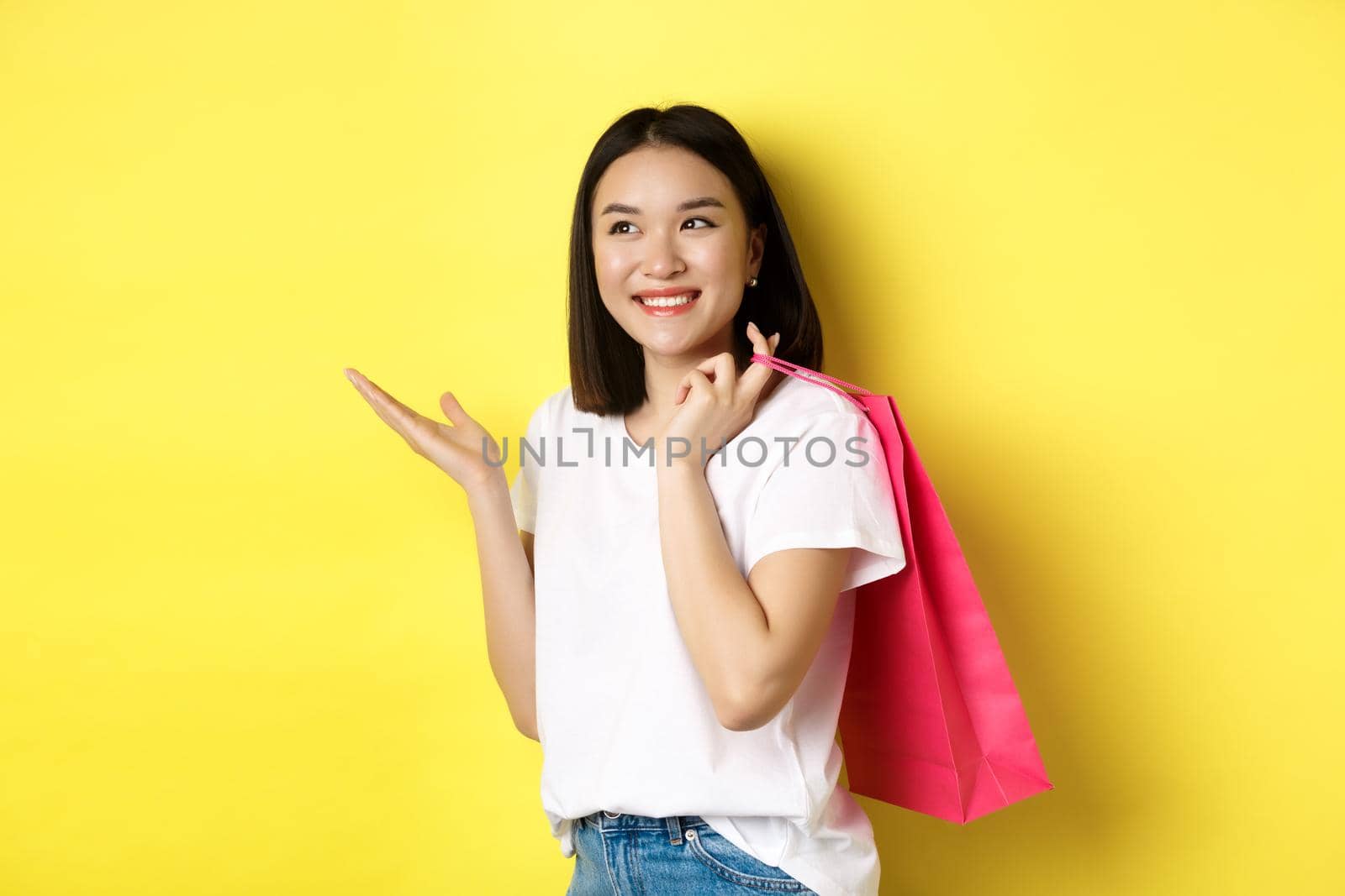 Cheerful asian female shopper looking amused, holding shopping bag and pointing hand left at store banner, standing over yellow background.