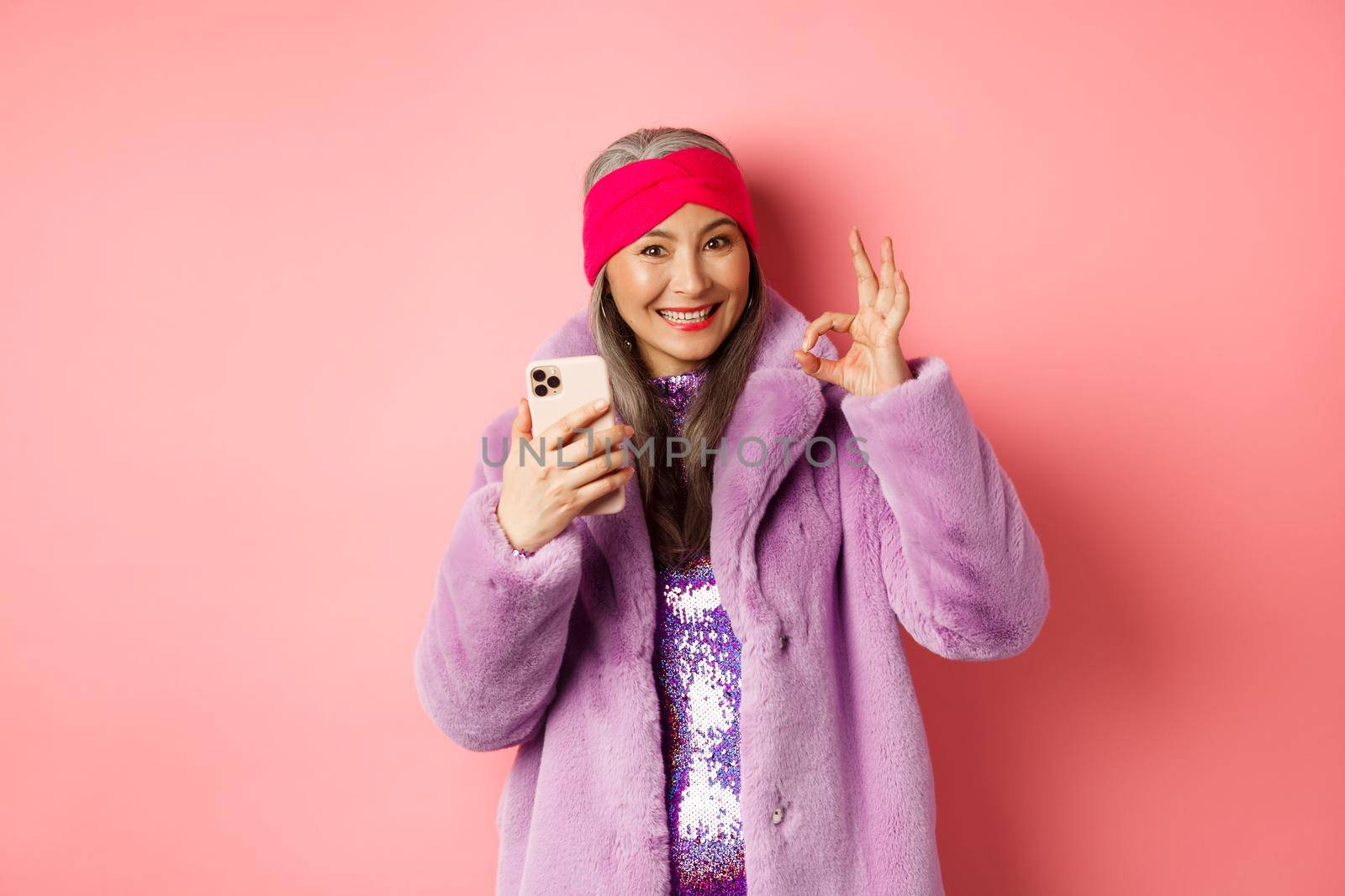 Online shopping and fashion concept. Stylish asian senior woman showing okay sign and holding mobile phone, recommending internet store, pink background.