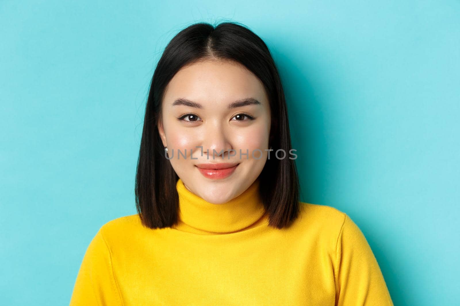 Beauty and skin care concept. Close up of beautiful young asian woman with perfect face, without blemishes, smiling at camera, standing over blue background.