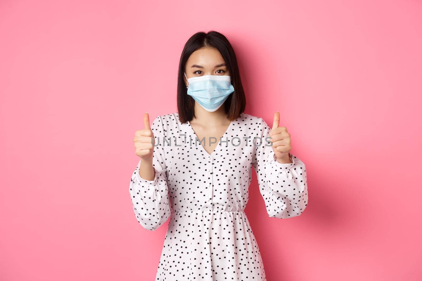 Covid-19, quarantine and lifestyle concept. Beautiful asian woman in dress and face mask showing thumbs-up, give approval, like and praise, standing over pink background by Benzoix