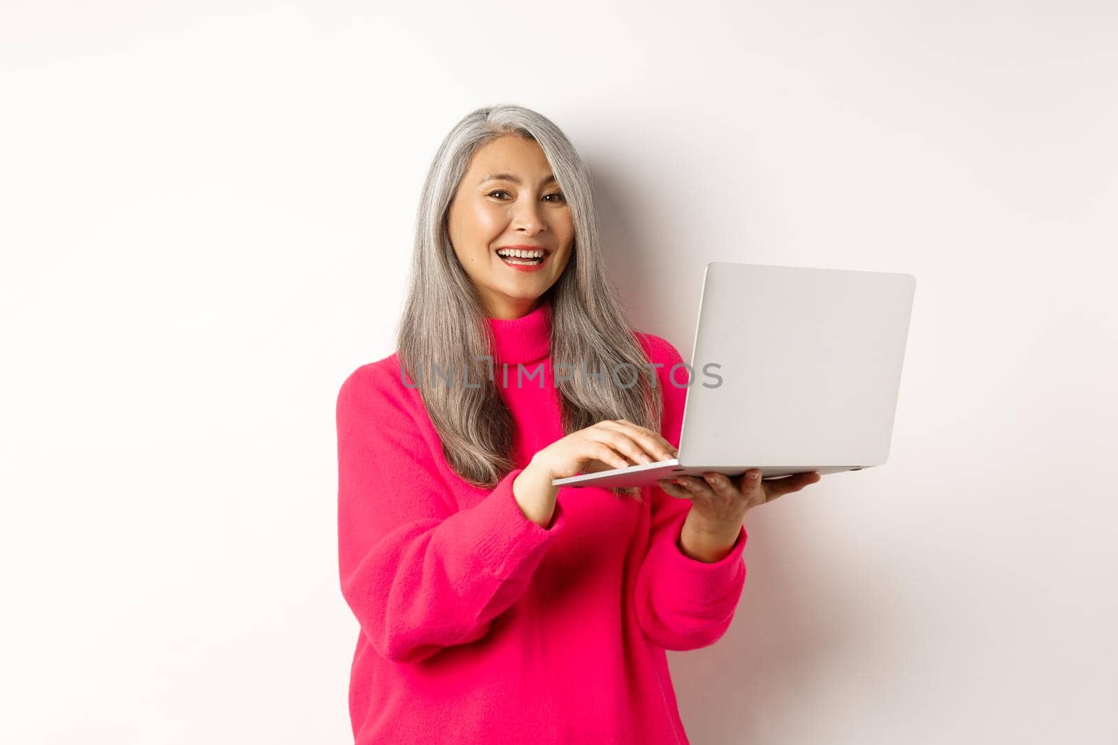 Beautiful asian senior woman entrepreneur working with laptop, laughing and smiling at camera, standing over white background.