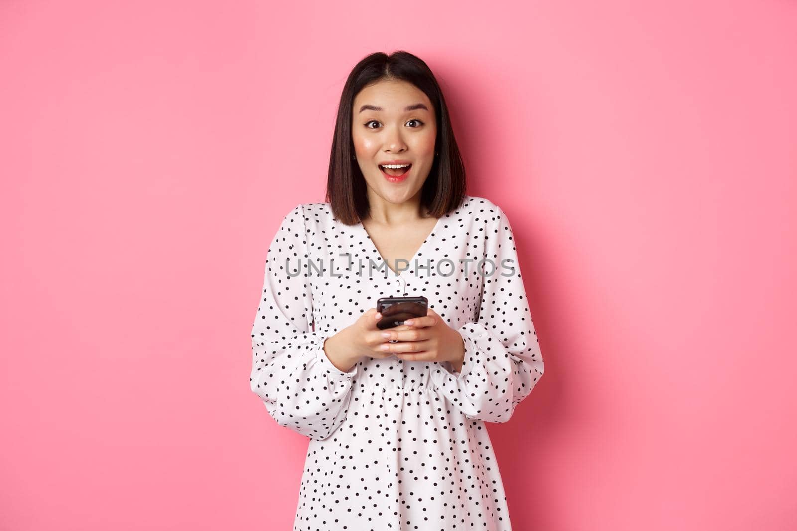 Online shopping. Amazed asian woman looking at camera with happy smile, making purchase with smartphone, using mobile phone app, standing over pink background.