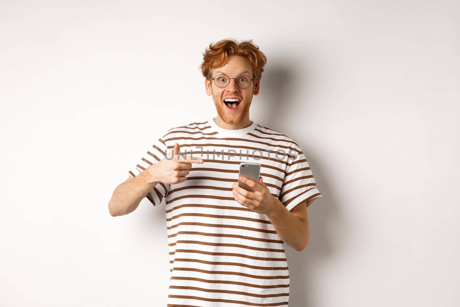 Technology and e-commerce concept. Amazed redhead man checking out online promo offer on mobile phone, pointing finger at smartphone and smiling, white background by Benzoix