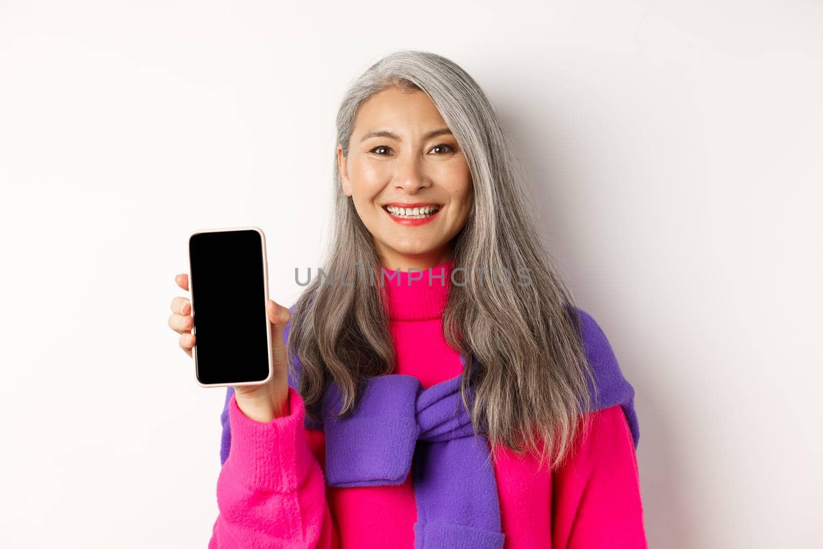 Online shopping. Close-up of smiling asian grandmother showing blank smartphone screen, recommending promotion, standing over white background.