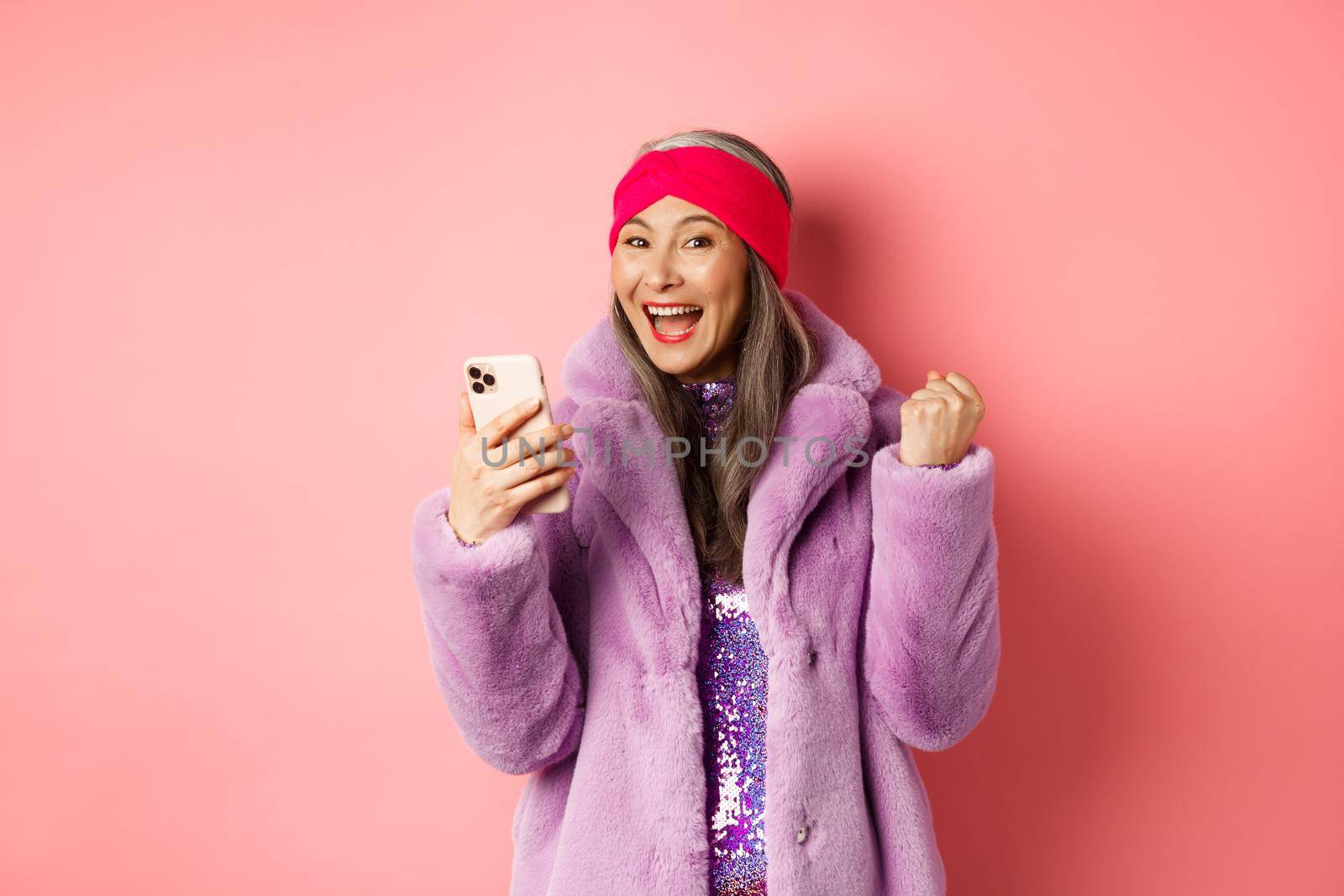 Online shopping and fashion concept. Happy asian senior woman winning prize in internet, holding mobile phone and making fist pump, scream of joy, standing over pink background.