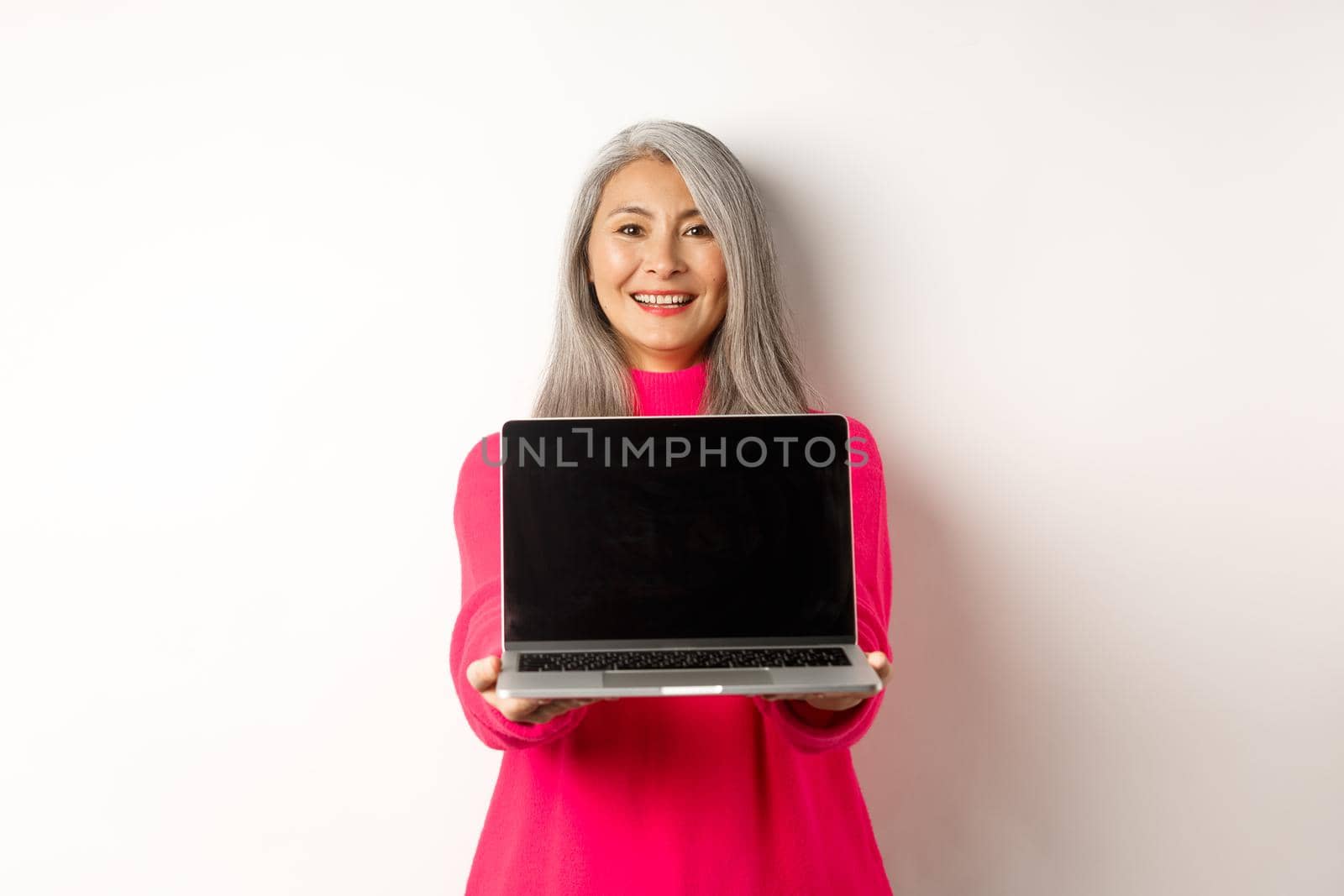 E-commerce concept. Smiling asian senior woman showing blank laptop screen and looking happy, demonstrating promo, white background.