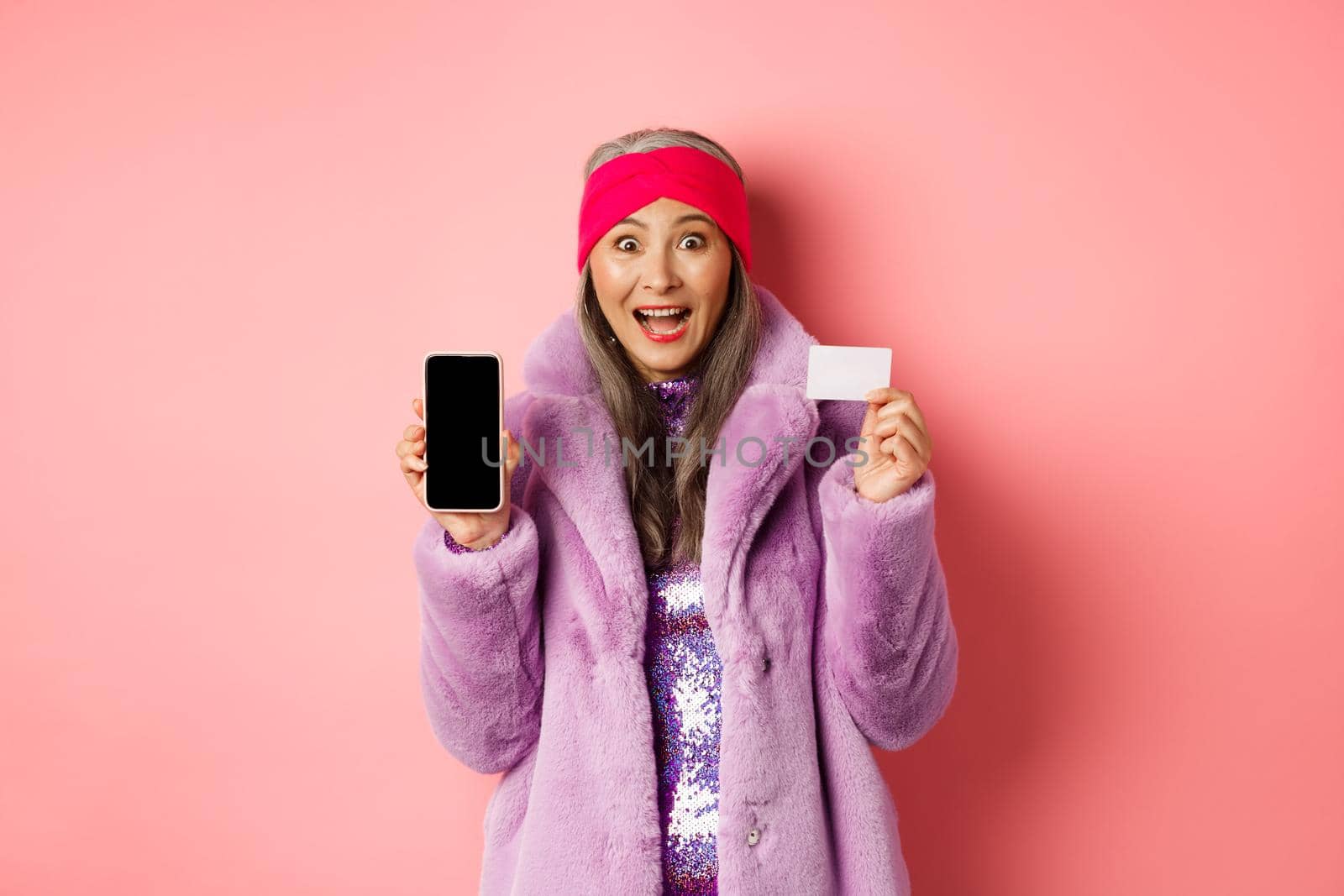 Special promotion. Excited asian senior woman showing blank smartphone screen and plastic credit card, checking out online offer, pink background.