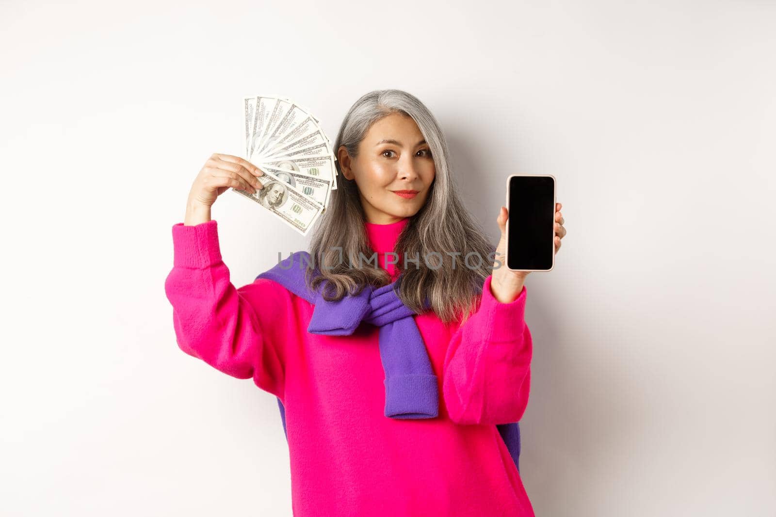 Fashionable asian senior woman showing money dollars and blank smartphone screen, demonstrate online shop, standing over white background.