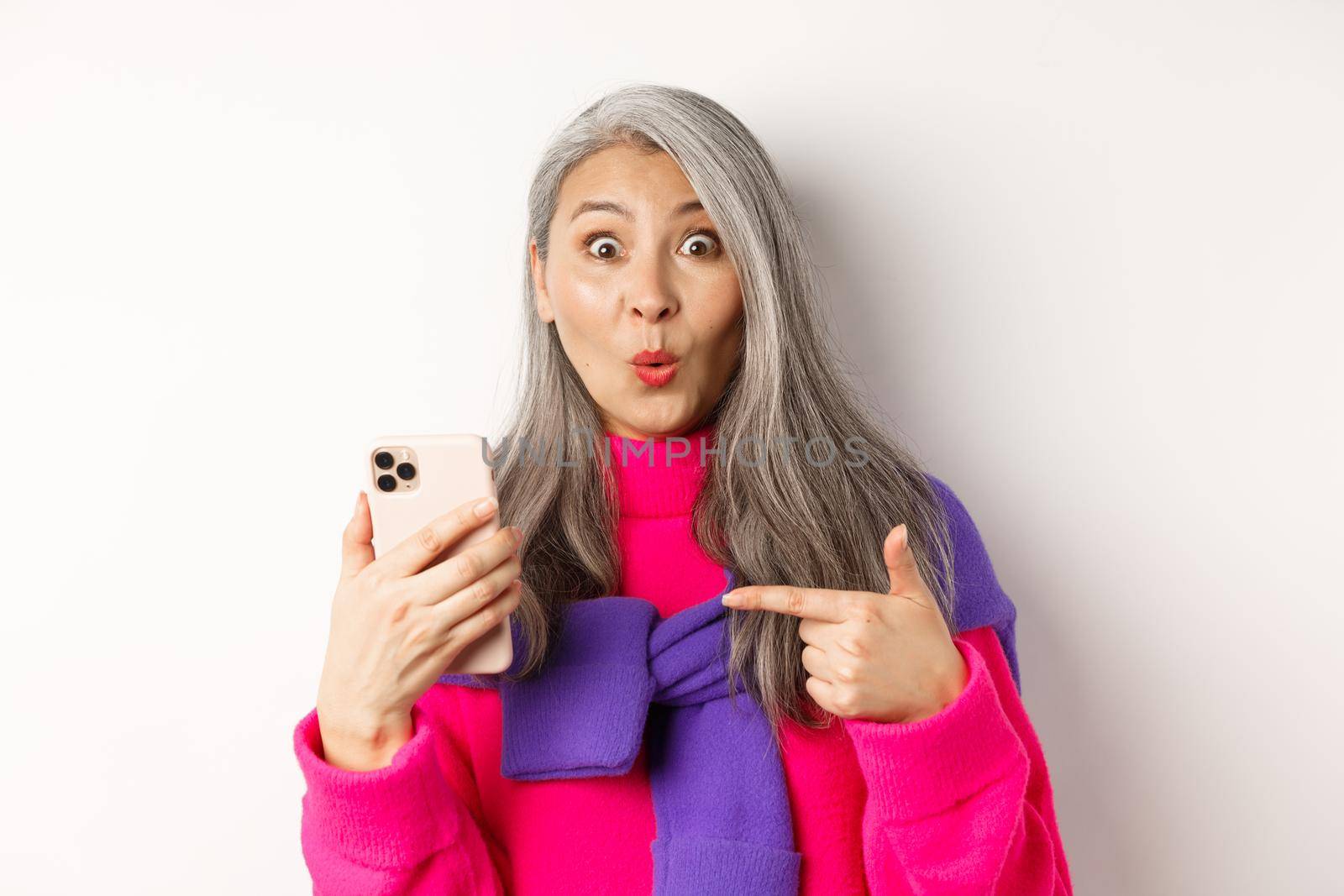 Online shopping. Close up of amazed asian senior woman pointing at smartphone and smiling amazed, standing over white background.