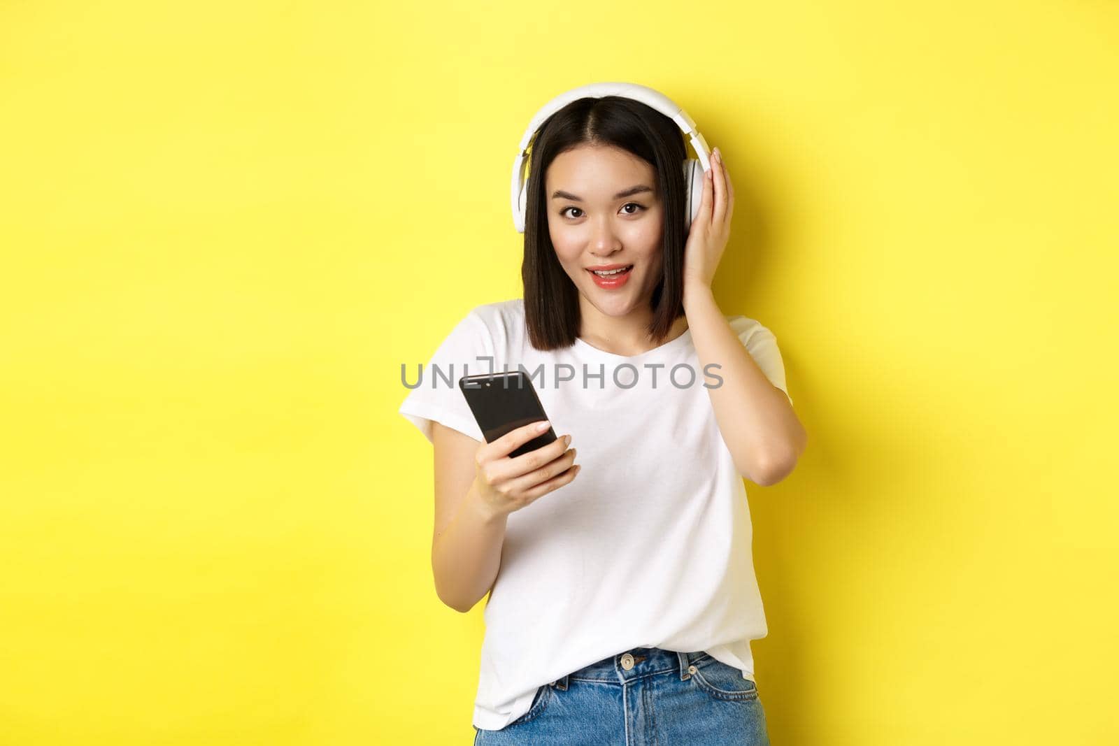 Modern asian woman listening music in wireless headphones, reading smartphone screen and smiling, standing in white t-shirt over yellow background.