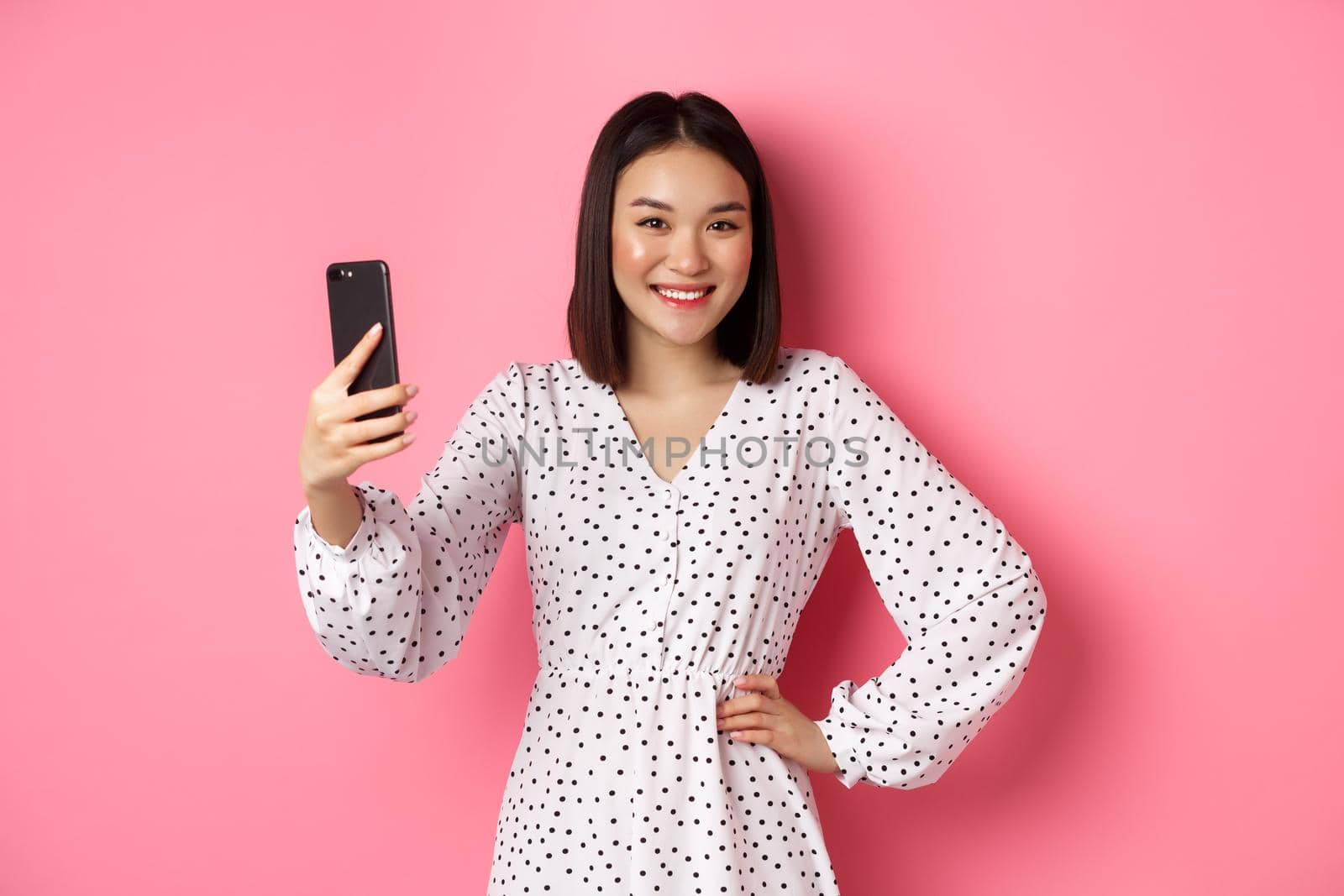 Beautiful asian girl using photo filters app and taking selfie on mobile phone, posing in cute dress against pink background.