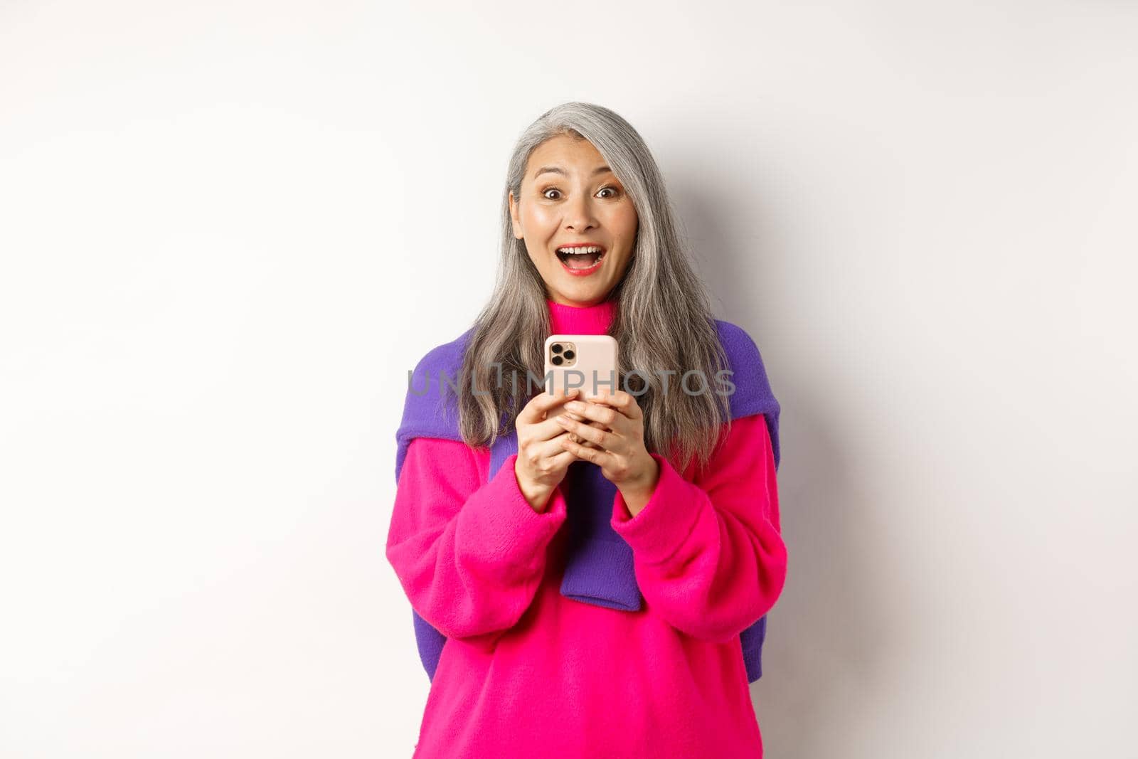 Surprised asian woman smiling at camera after reading promotion on smartphone, standing with mobile phone over white background.