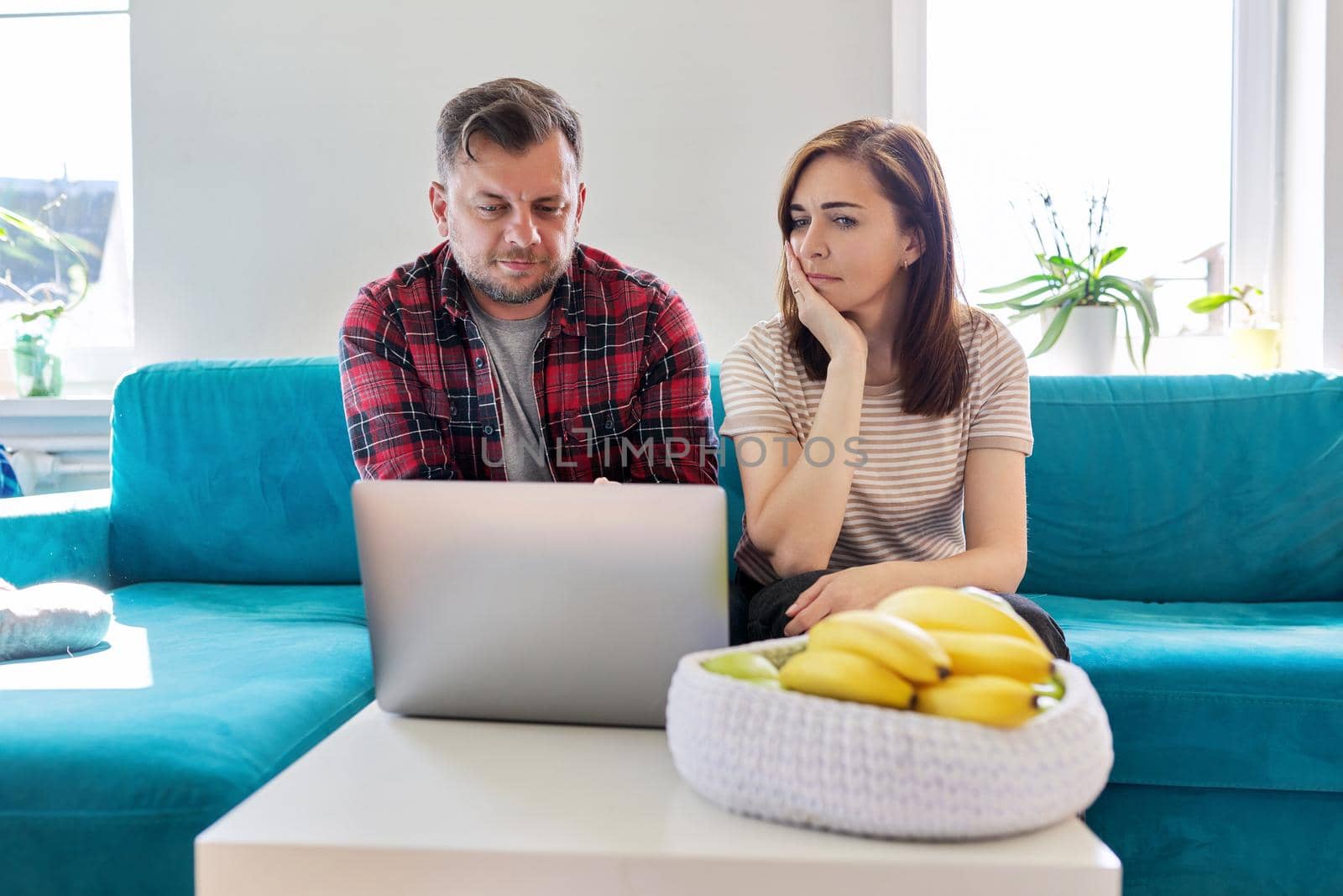 Serious middle aged couple looking at laptop screen at home in living room. Concentrated husband and wife listening looking into laptop, online information. Family, technology, lifestyle, 40s people