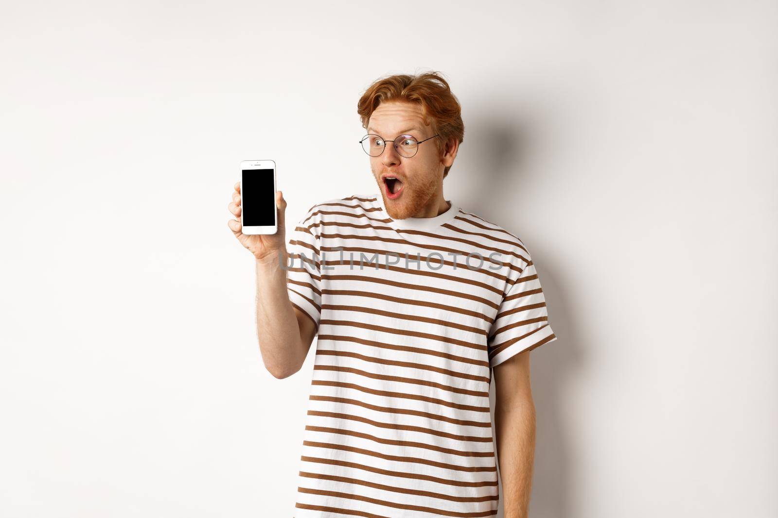 Technology and e-commerce concept. Surprised and shocked redhead guy checking out online promotion, showing blank smartphone screen and drop jaw, white background by Benzoix