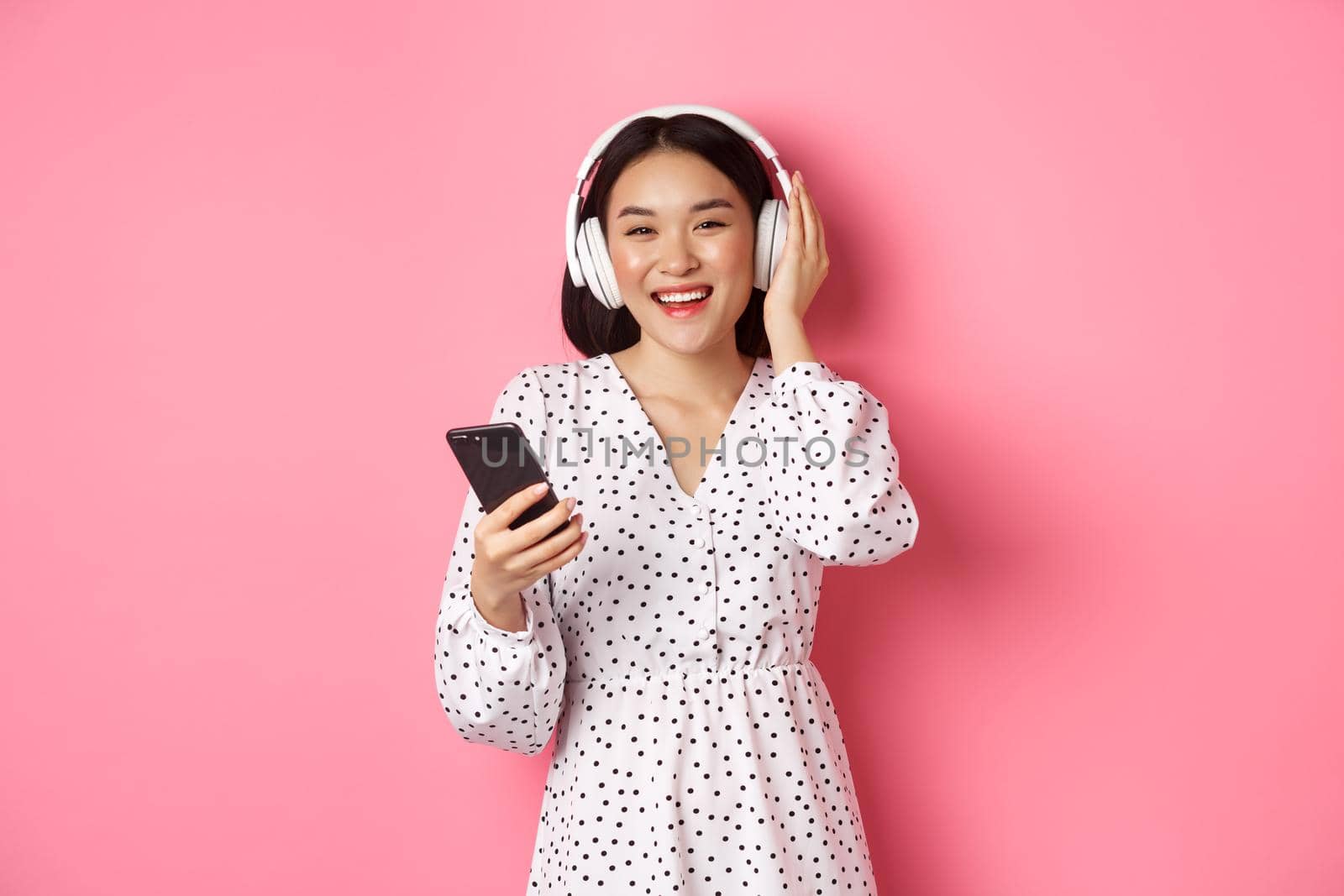 Beautiful asian woman listening music in headphones, using mobile phone, smiling happy at camera, standing over pink background.