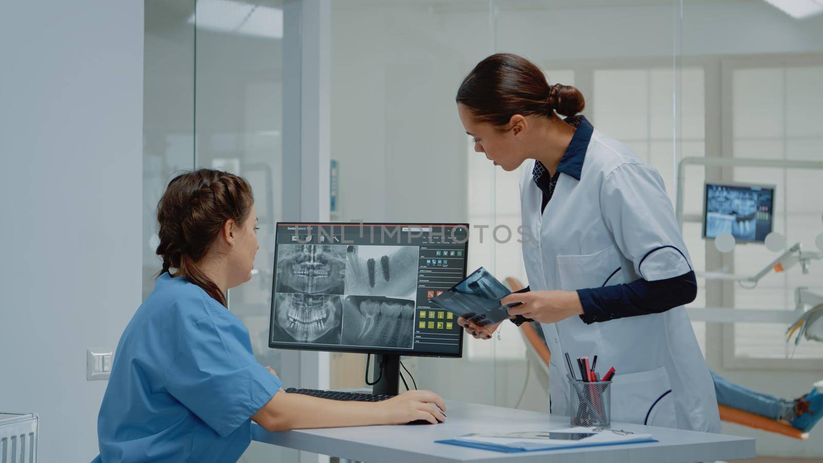 Dentist holding dentition x ray scan comparing to radiography on computer monitor at dental clinic. Nurse sitting at desk looking at screen for teeth examination and consultation