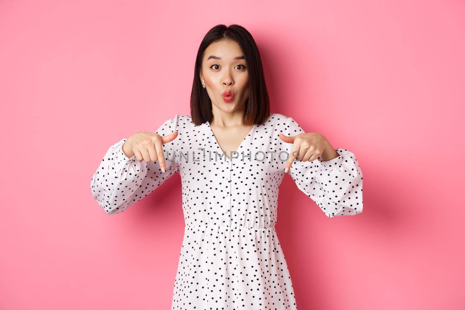 Impressed asian woman in dress pointing fingers down, look there gesture, check out discounts and sales, standing amazed over pink background.