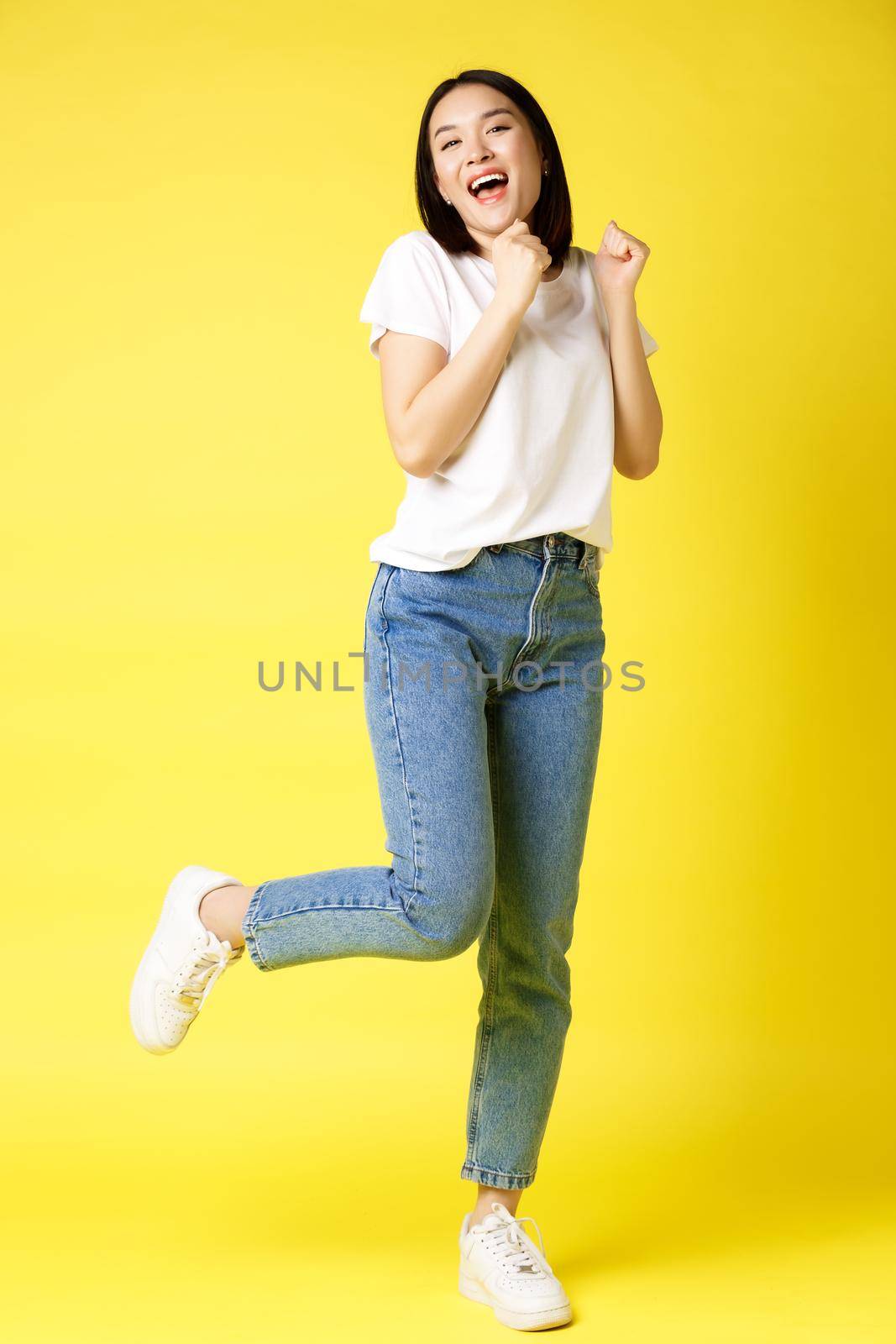 Full size shot of happy asian woman dancing and jumping from happiness, winning and celebrating victory, posing over yellow background by Benzoix