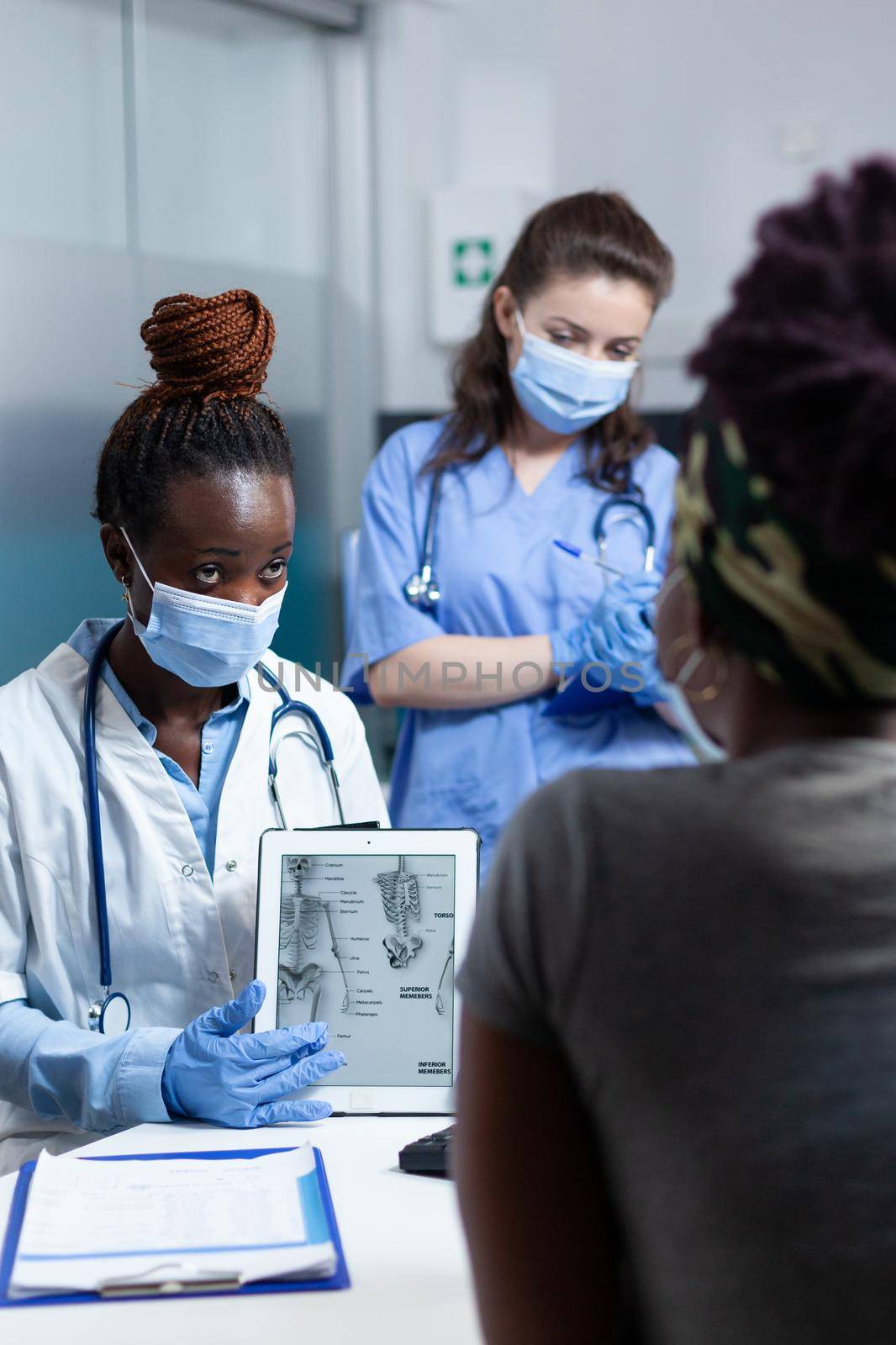 African american doctor with face mask against coronavirus explaining bones radiography to sick patient during clinical appointment in hospital office. Radiologist nurse writing medication treatment