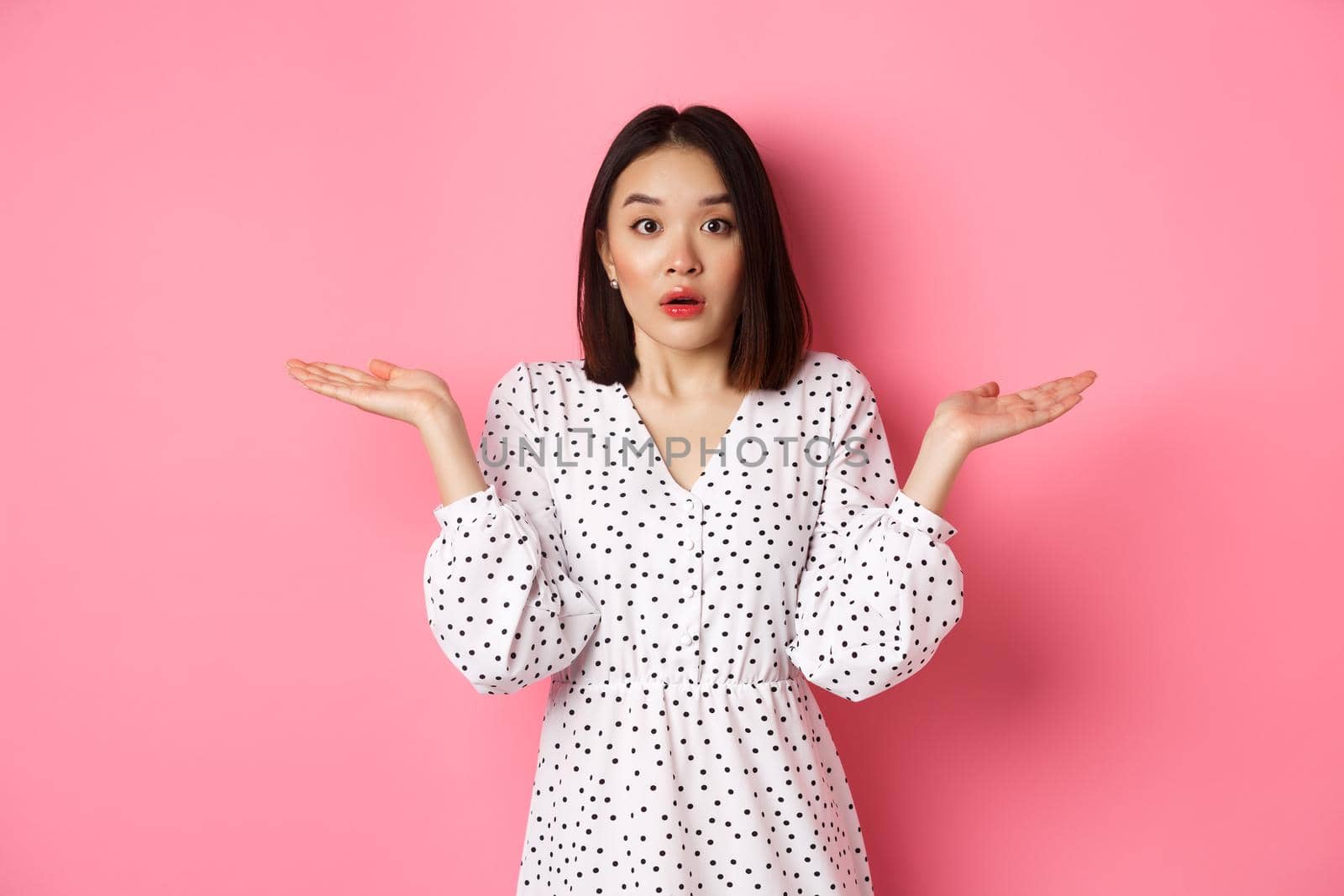 Confused and clueless asian girl shrugging, staring at camera puzzled, dont know what to do, spread hands sideways, standing over pink background.