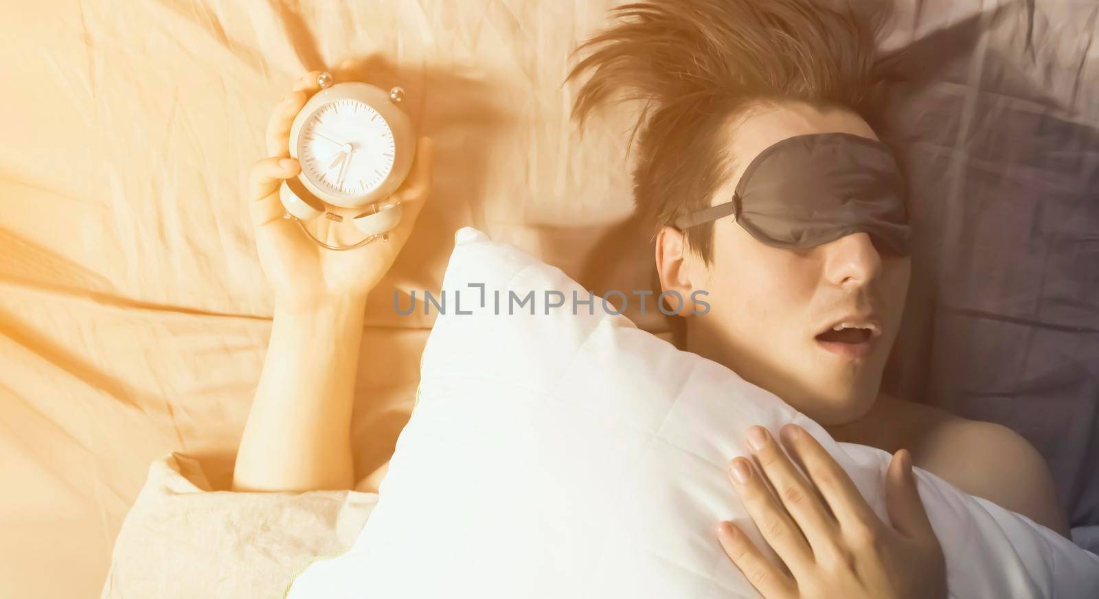 A young funny man sleeps soundly early in the morning, hugs a pillow and holds a vintage alarm clock in his hands, the sleep mask has slipped from his eyes.