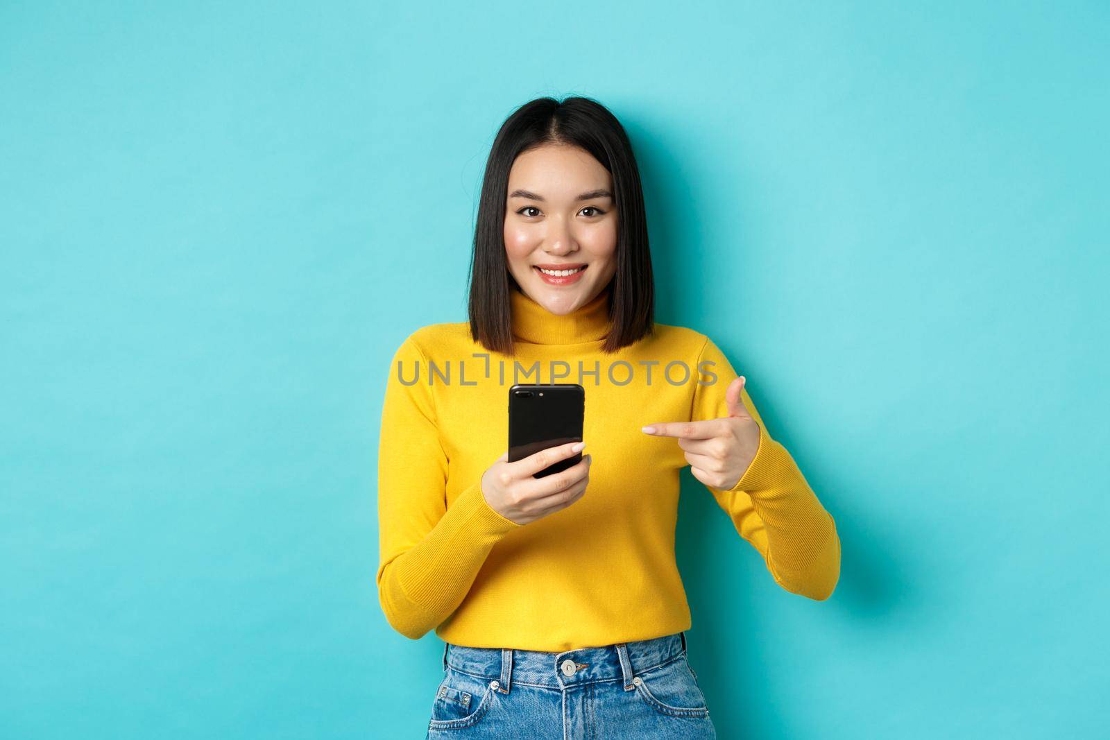 E-commerce and online shopping concept. Cute asian woman in yellow sweater pointing at smartphone, smiling at camera, standing over blue background.