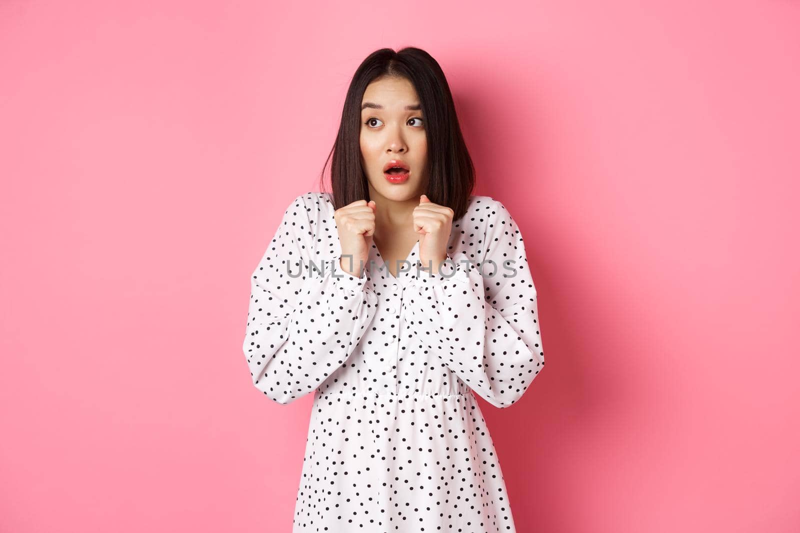 Scared and timid asian girl trembling from fear, staring left and gasping, standing in dress over pink background.