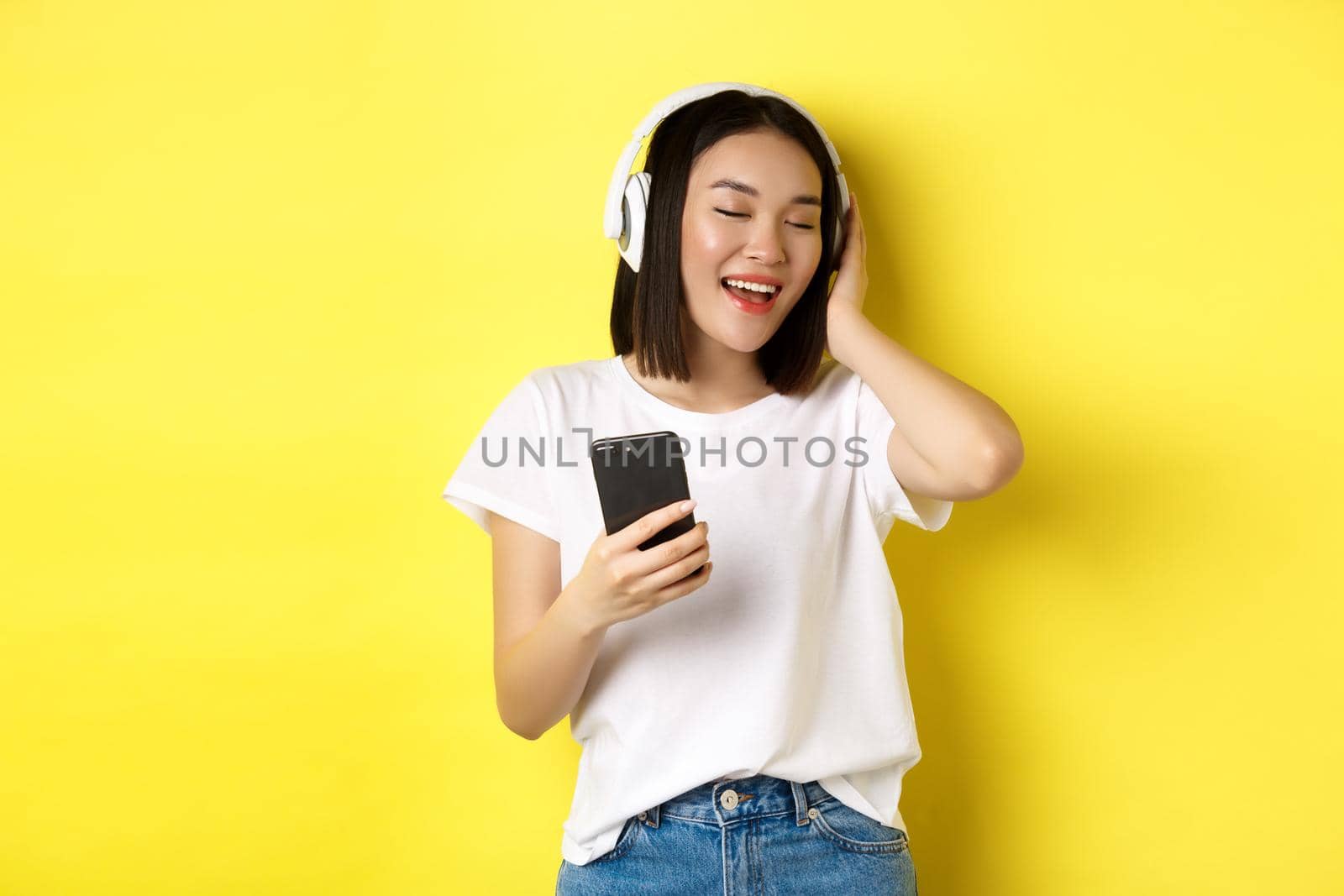 Cool asian girl singing along and listening music in wireless headphones, holding smartphone in hand, standing over yellow background.