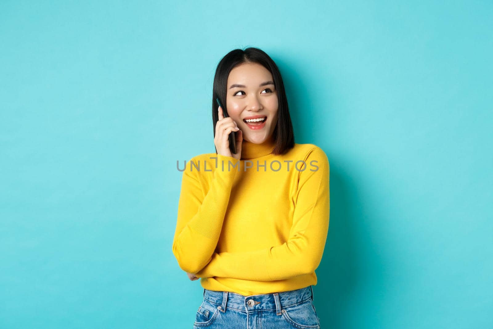 Stylish korean woman calling on phone, talking on smartphone and looking happy at upper right corner, smiling carefree, standing in relaxed pose over blue background.