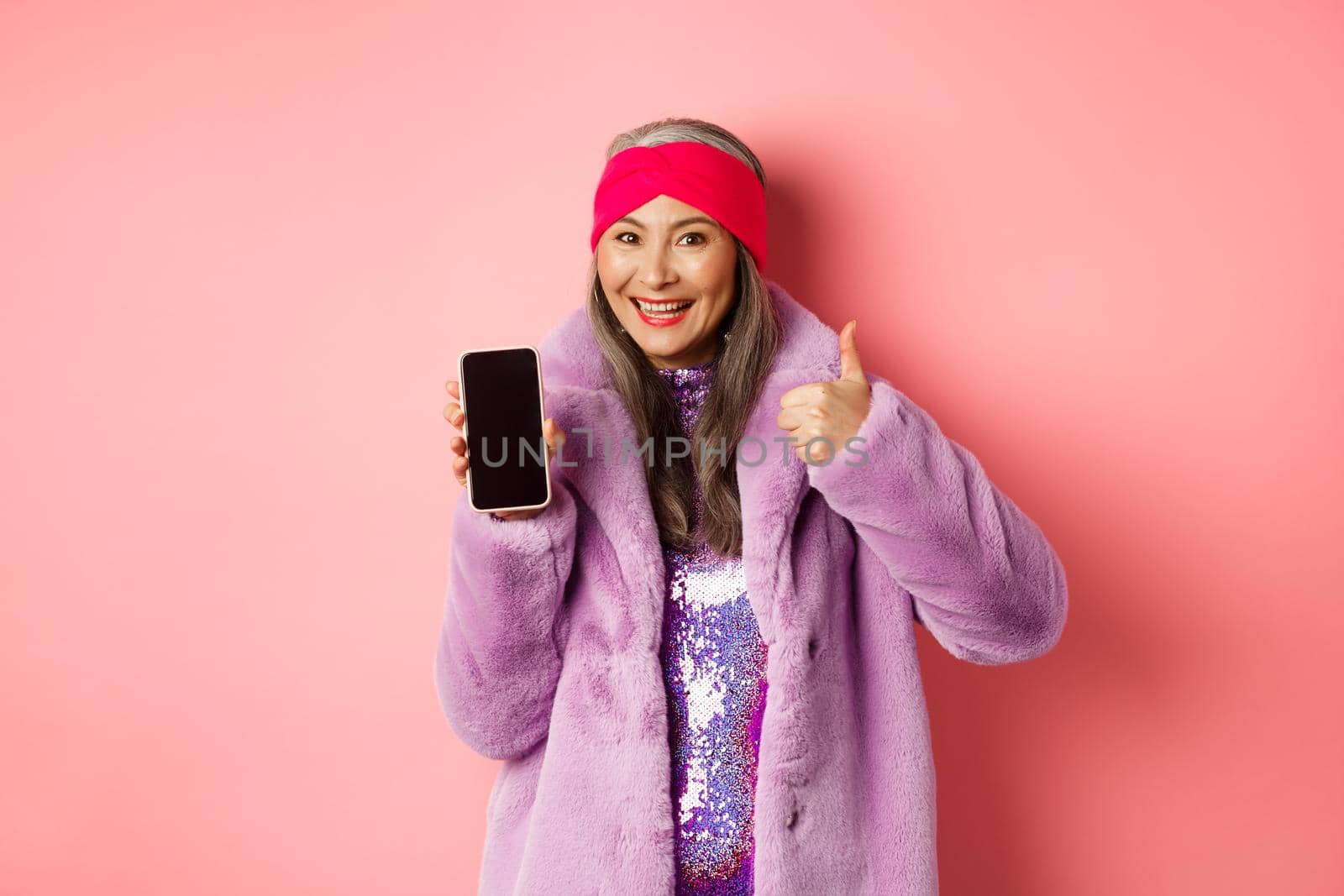 Online shopping and fashion concept. Funky asian senior woman showing blank mobile screen and thumbs-up, like and recommend internet promo, pink background.