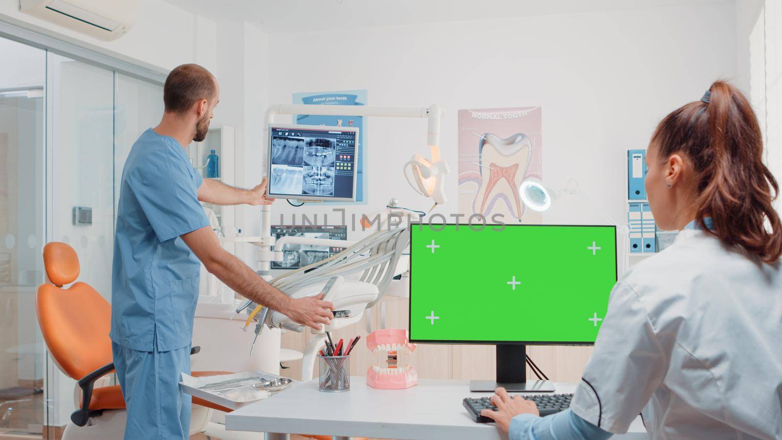 Woman working as dentist with green screen on monitor at dental cabinet while assistant preparing tools for teethcare. Specialist using computer with chroma key and mockup template