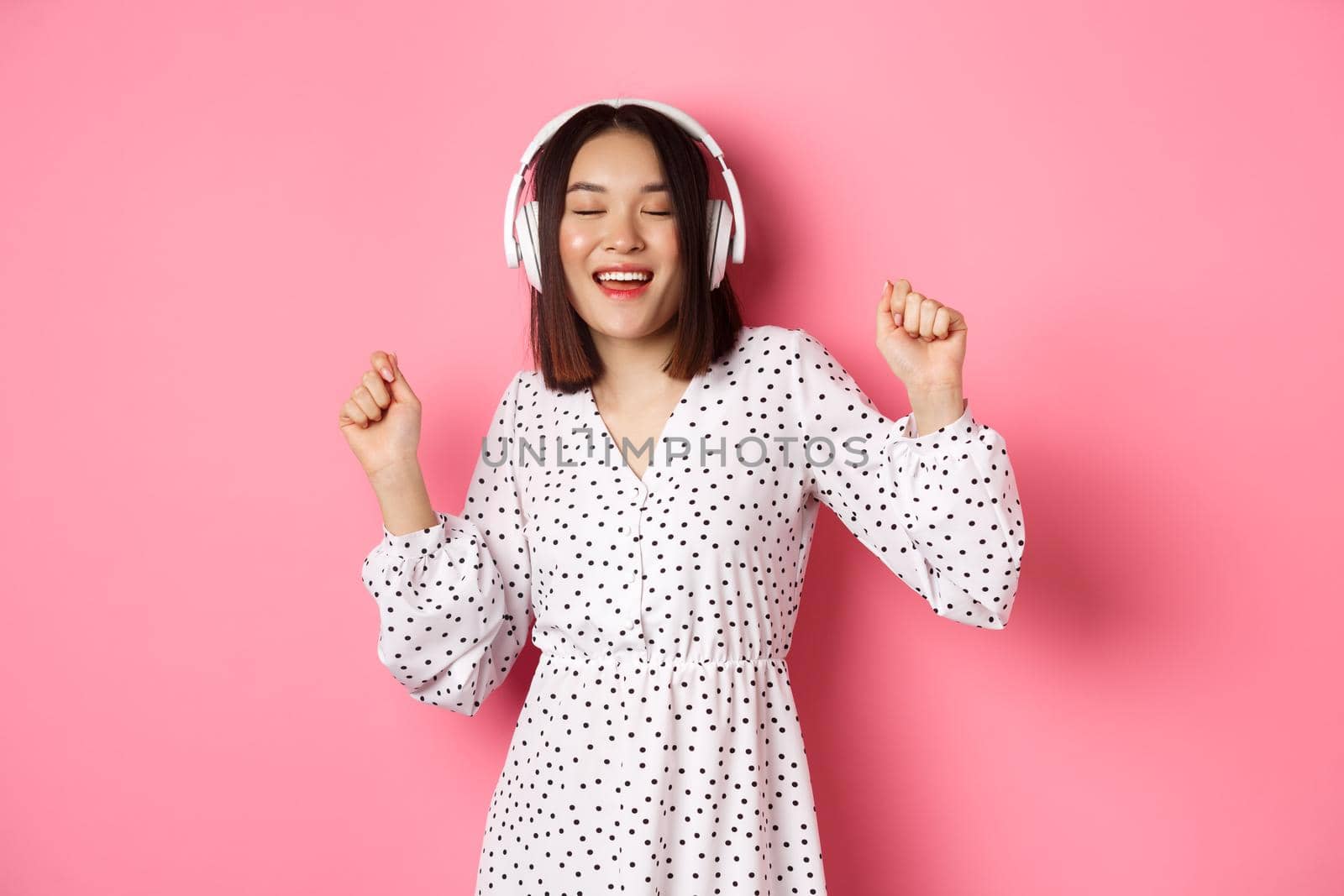 Happy young asian woman dancing and having fun, listening music in headphones, standing over pink background. Copy space