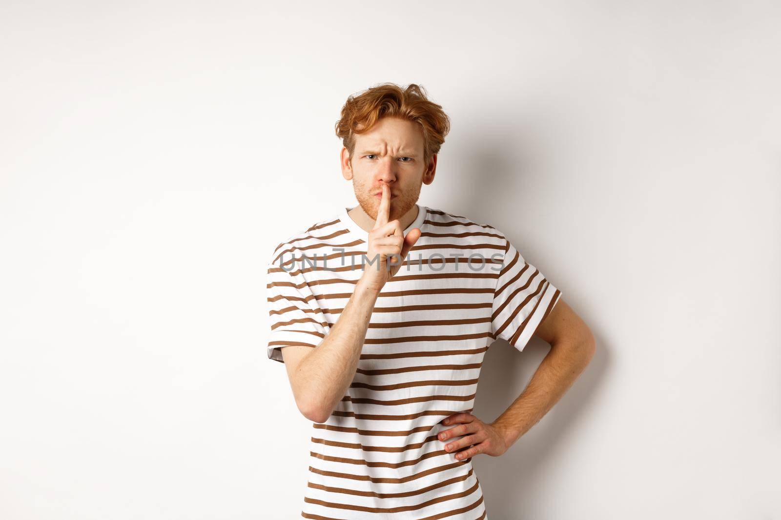 Angry redhead man looking displeased and hushing at camera, making shh gesture, standing over white background.