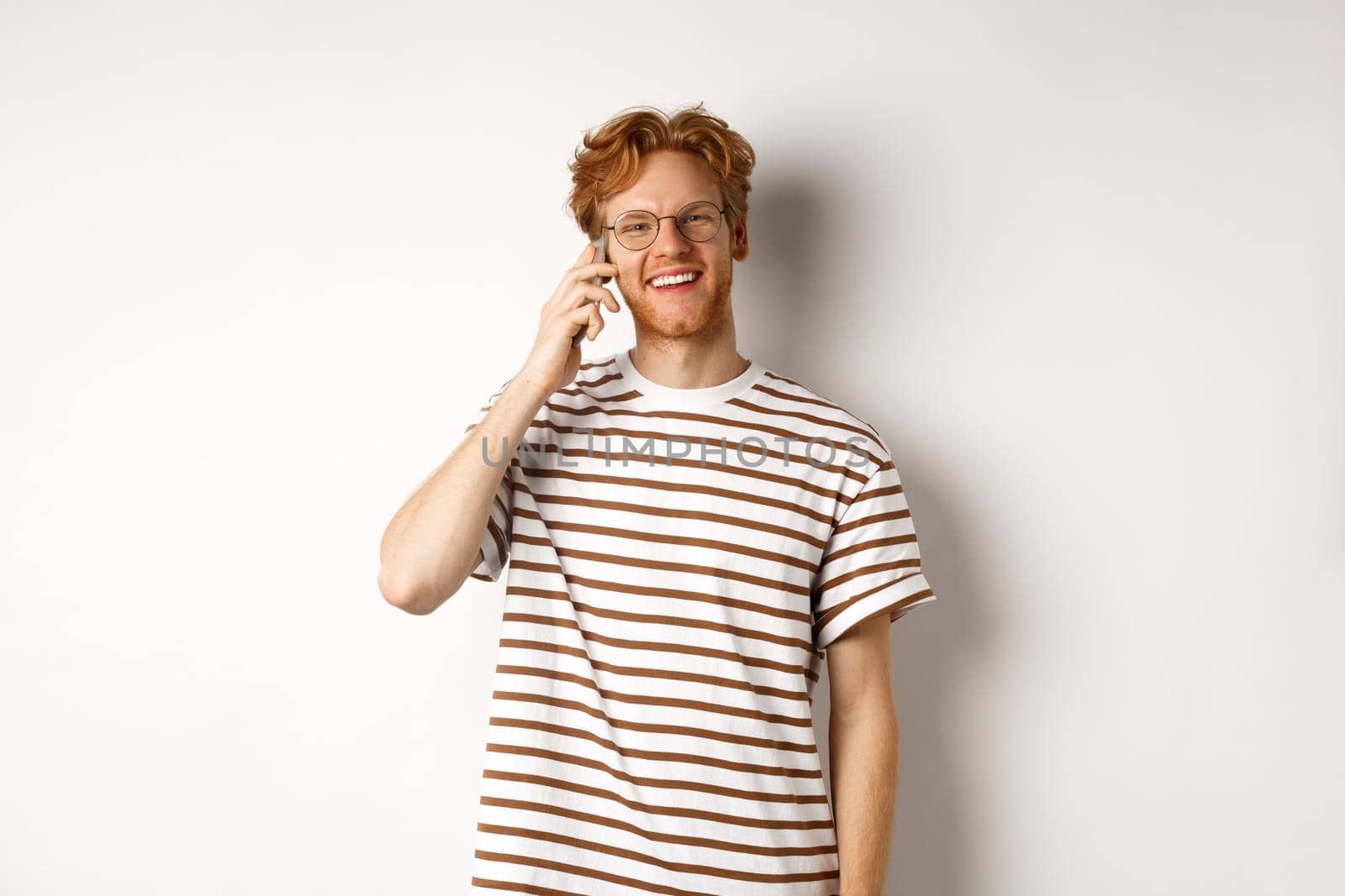 Hipster with red hair and glasses talking on mobile phone, smiling during conversation, white background.