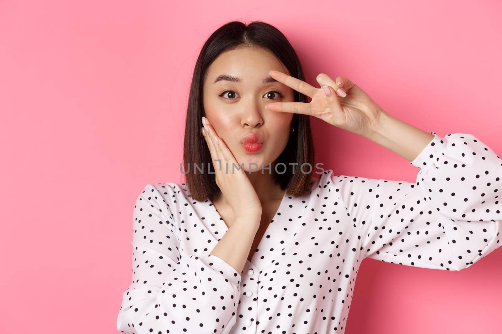 Beauty and lifestyle concept. Close-up of pretty asian woman showing peace sign and touching cheek, smiling happy at camera, standing over pink background.