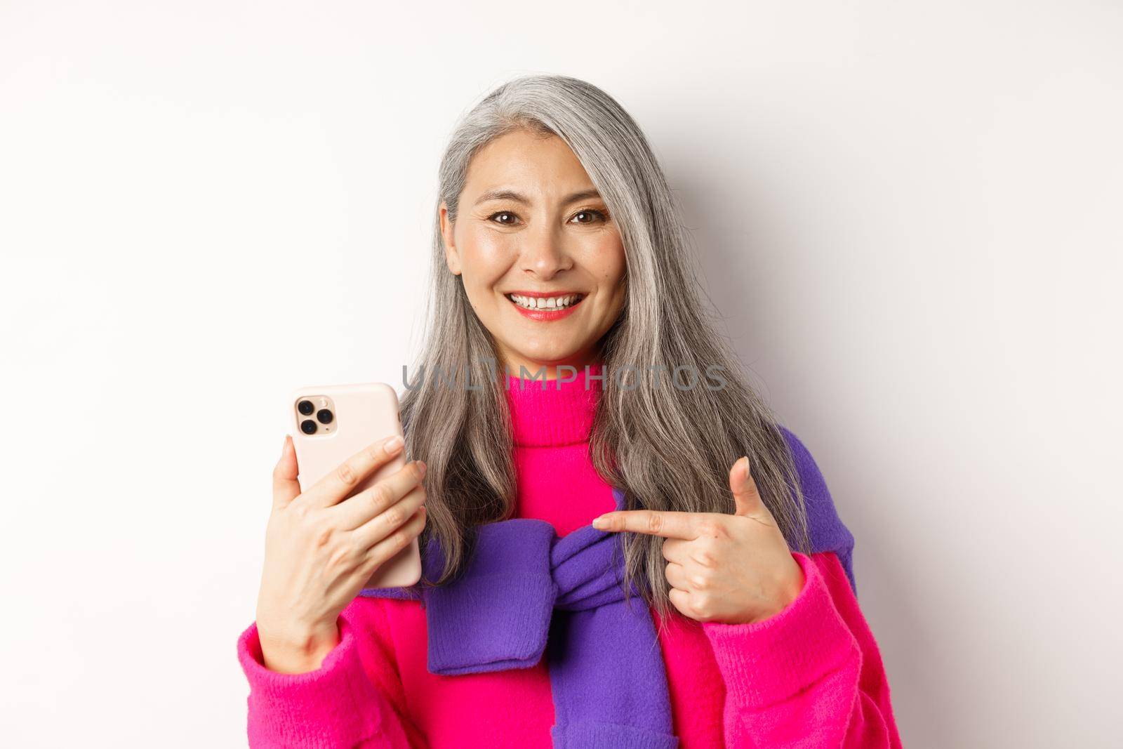 Close up of pretty asian senior woman with grey hair, pointing finger at smartphone and smiling, recommending mobile app, standing over white background.