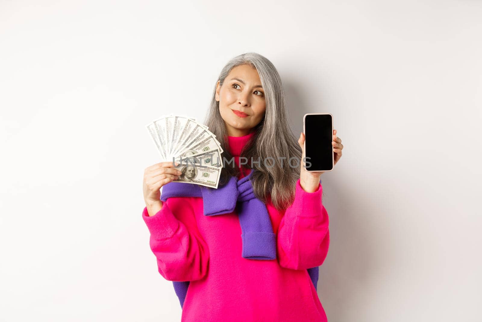 Fashionable asian senior woman showing money dollars and blank smartphone screen, demonstrate online shop, standing over white background.