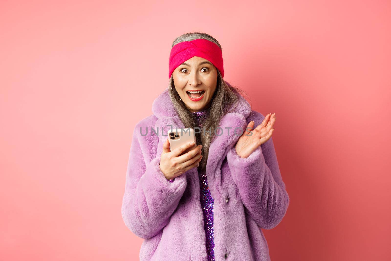 Online shopping and fashion concept. Asian mature woman looking excited as checking out internet promo offer on smartphone, looking happy at camera, pink background.