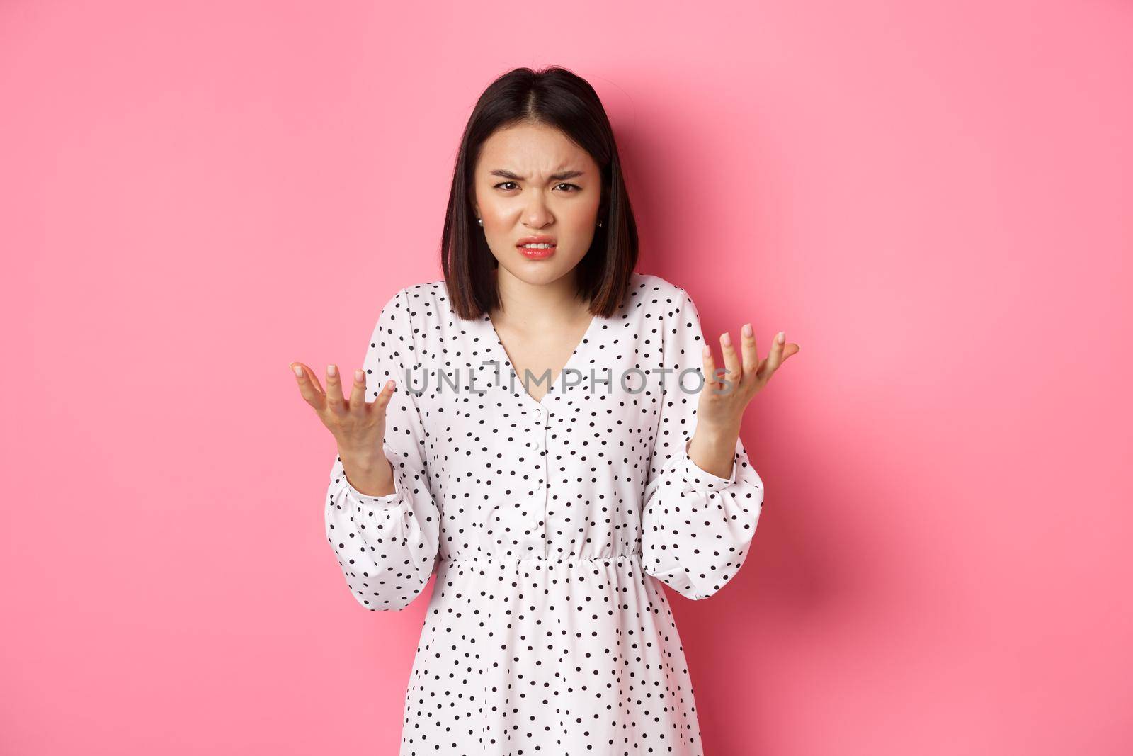 Angry and confused asian woman pointing hands at camera and grimacing furious, standing annoyed against pink background by Benzoix