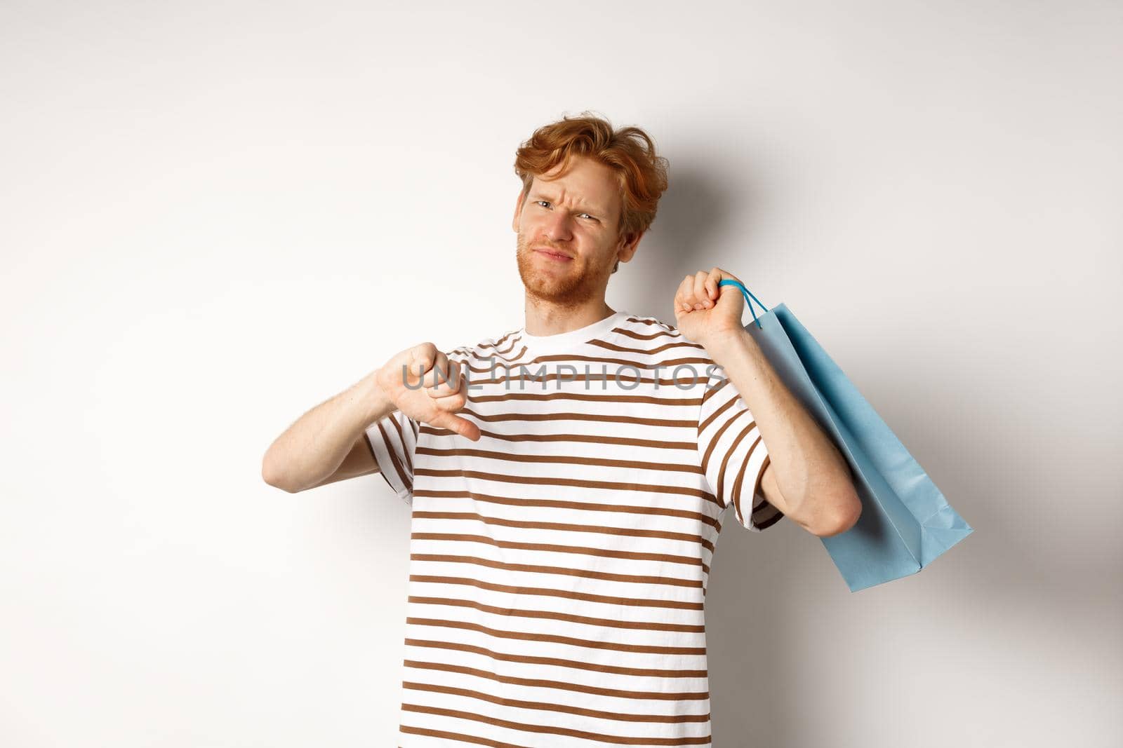 Disappointed young man with red hair and beard showing thumbs-down after bad shopping experience, holding bag over shoulder and frowning upset, white background by Benzoix