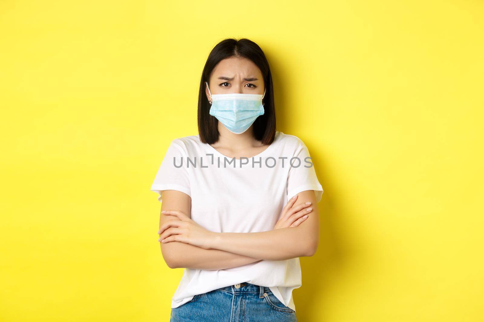 Covid, health care and pandemic concept. Asian woman in white t-shirt and medical mask cross arms on chest and looking sad and disappointed at camera.