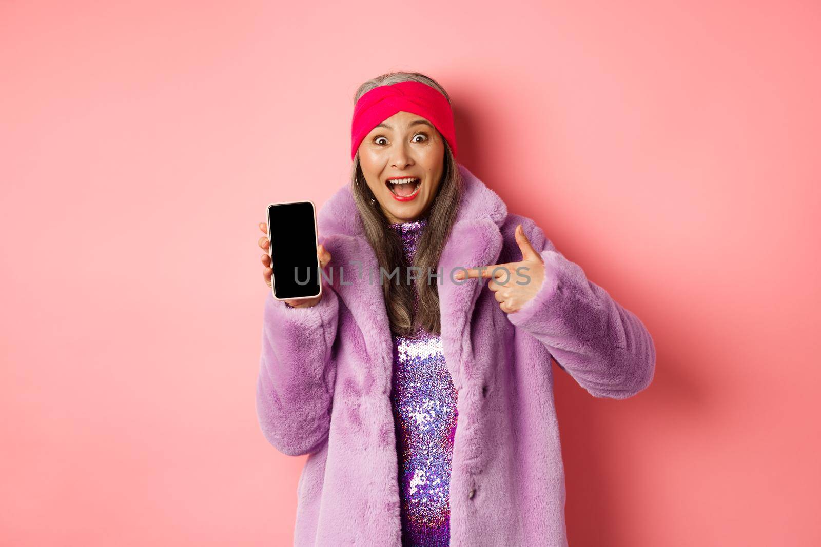 Online shopping and fashion concept. Stylish asian senior woman in faux fur coat pointing finger at blank smartphone screen, smiling happy, showing application on phone.