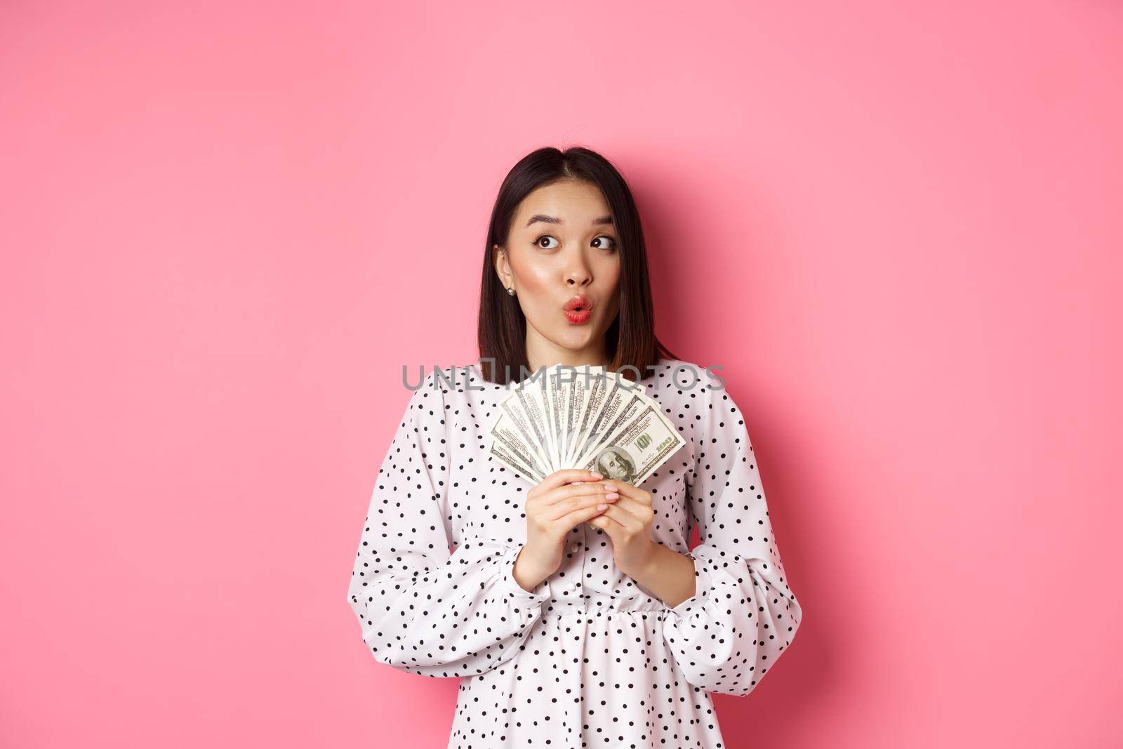 Shopping concept. Dreamy asian woman thinking, holding money dollars and looking aside thoughtful, standing over pink background.